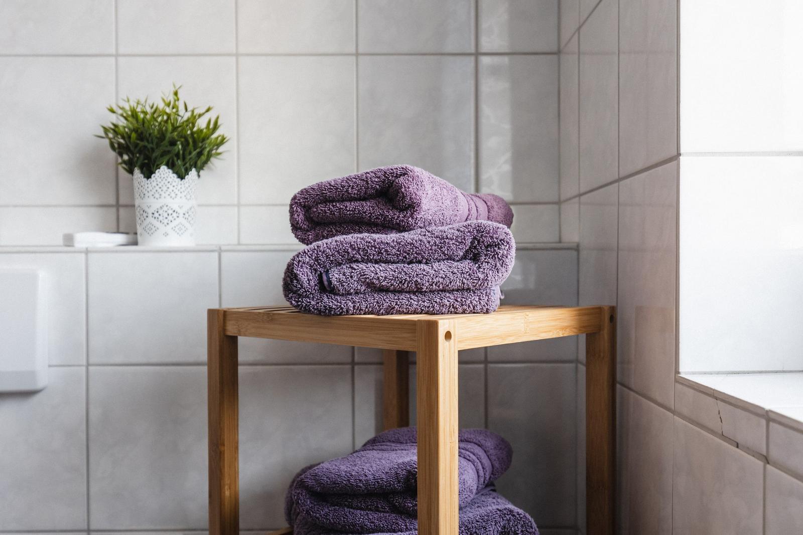 Dusche mit Holzregal, lila Handtücher und Pflanze auf weißer Fliesenwand.