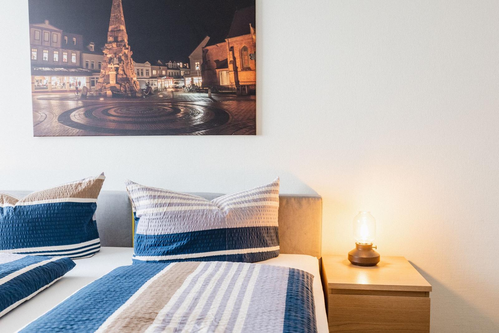 Schlafzimmer mit Bett, Nachttisch und Wandbild eines Platzes