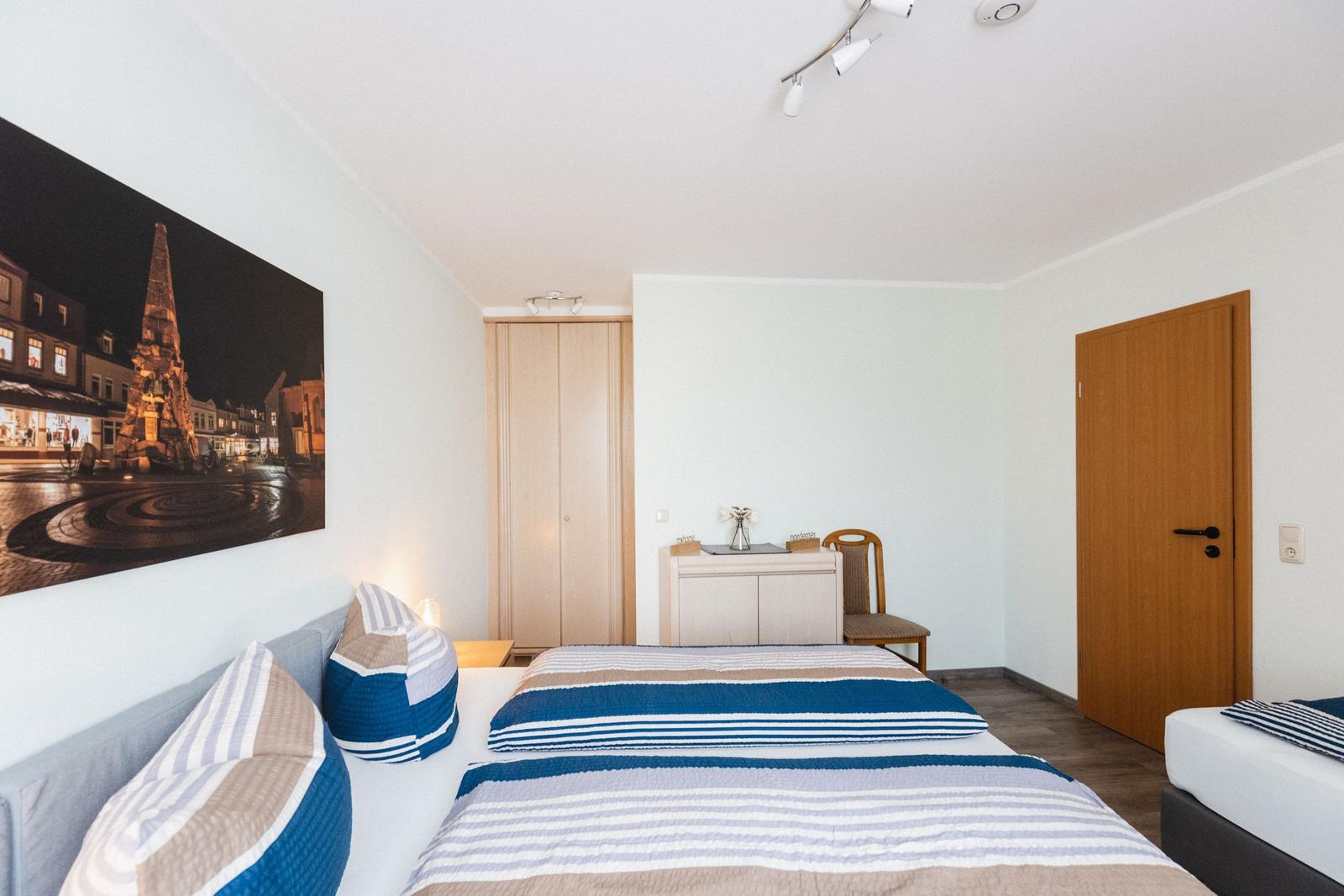 Schlafzimmer mit Bett, Kleiderschrank und Wandbild. Tür und Stuhl sind sichtbar.