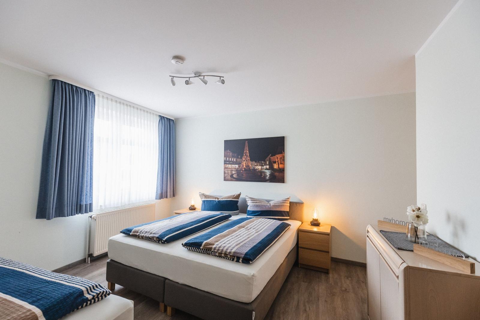 Schlafzimmer mit Doppelbett, blauen Vorhängen und Wandbild. Nebenbei steht ein Schrank und Nachttische.