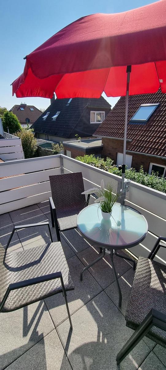Terrasse mit Tisch, Stühlen und rotem Sonnenschirm. Blick auf Dächer und Grünflächen.