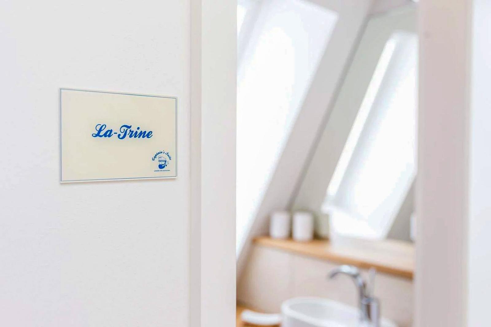 Schild mit Namen La-Trine an der Wand. Im Hintergrund ist ein Badezimmer mit Waschbecken und Spiegel sichtbar.