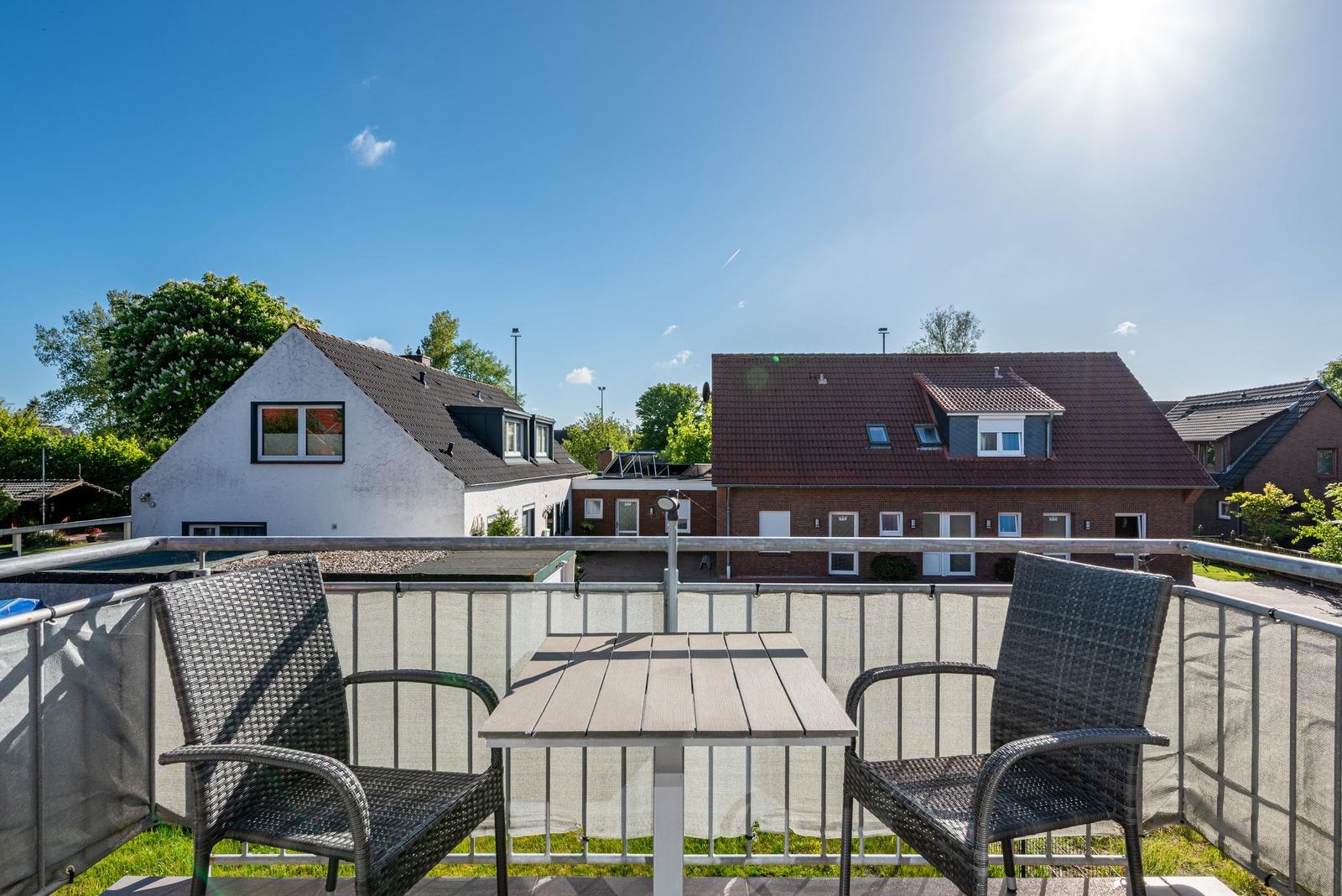 Terrasse mit Tisch und zwei Stühlen. Blick auf Wohnhäuser und Bäume unter blauem Himmel.