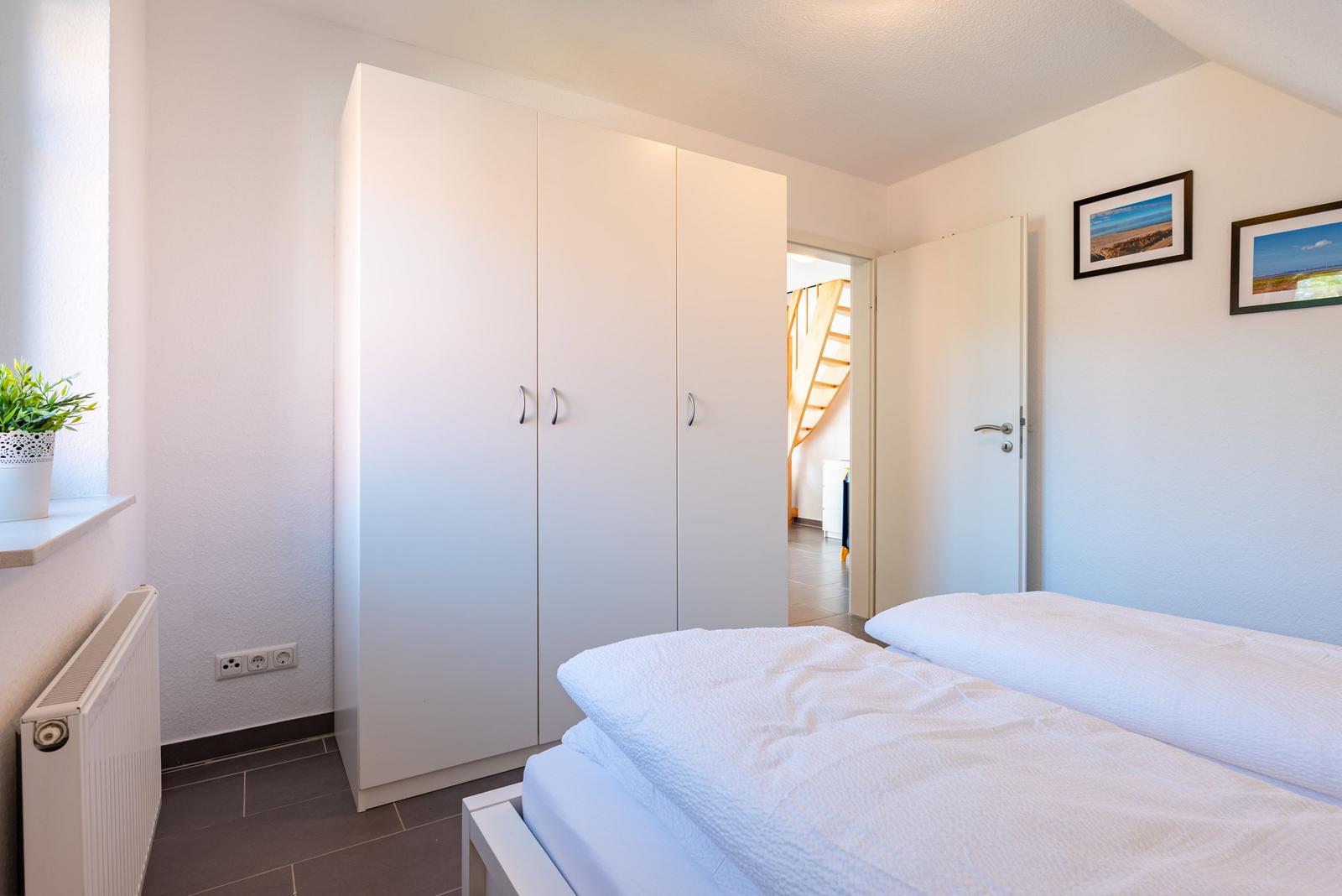 Schlafzimmer mit weißem Bett, Kleiderschrank und Fenster mit Pflanze.