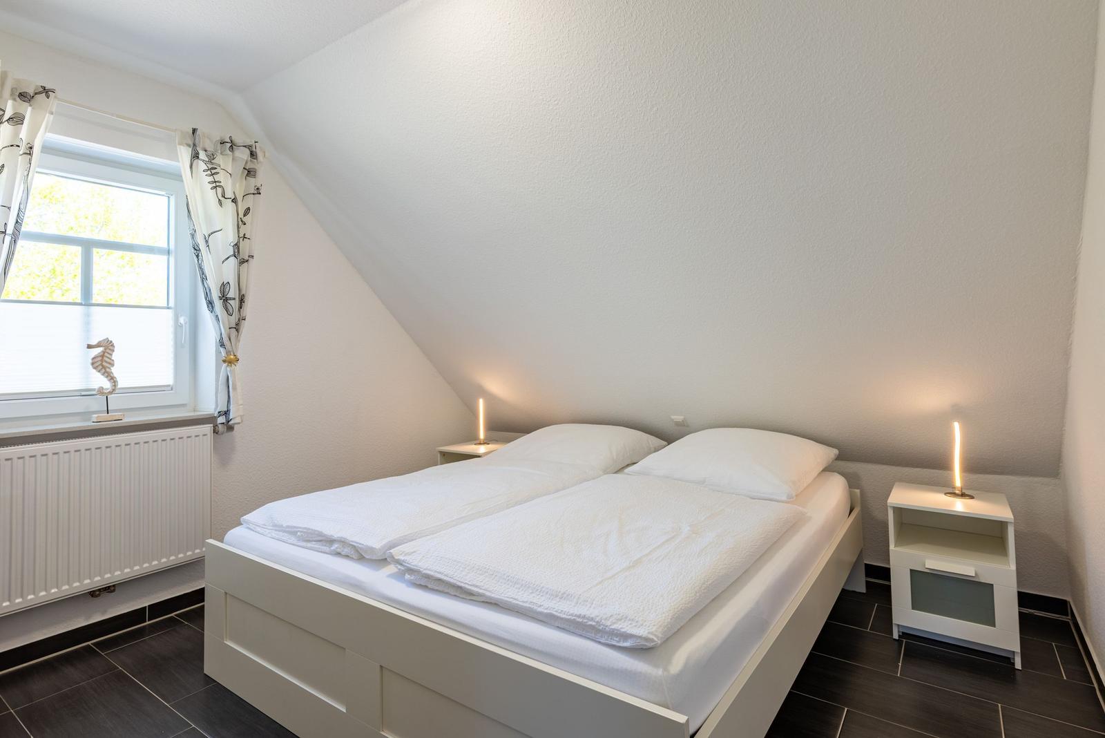 Doppelbett mit weißen Laken, zwei Nachttische mit Lampen und Fenster mit Vorhängen.