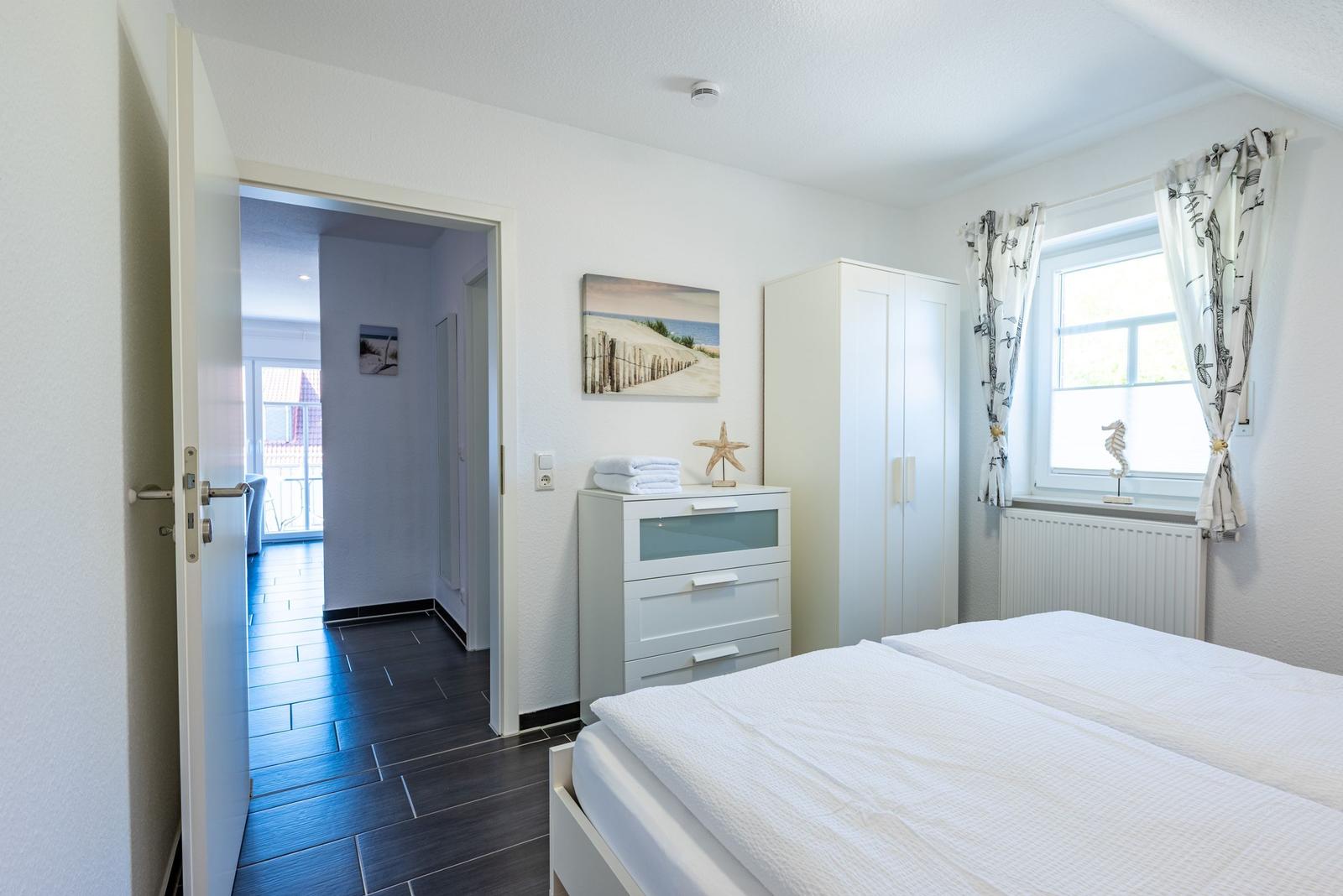 Schlafzimmer mit weißem Bett, Kleiderschrank und Fenster mit Vorhängen.
