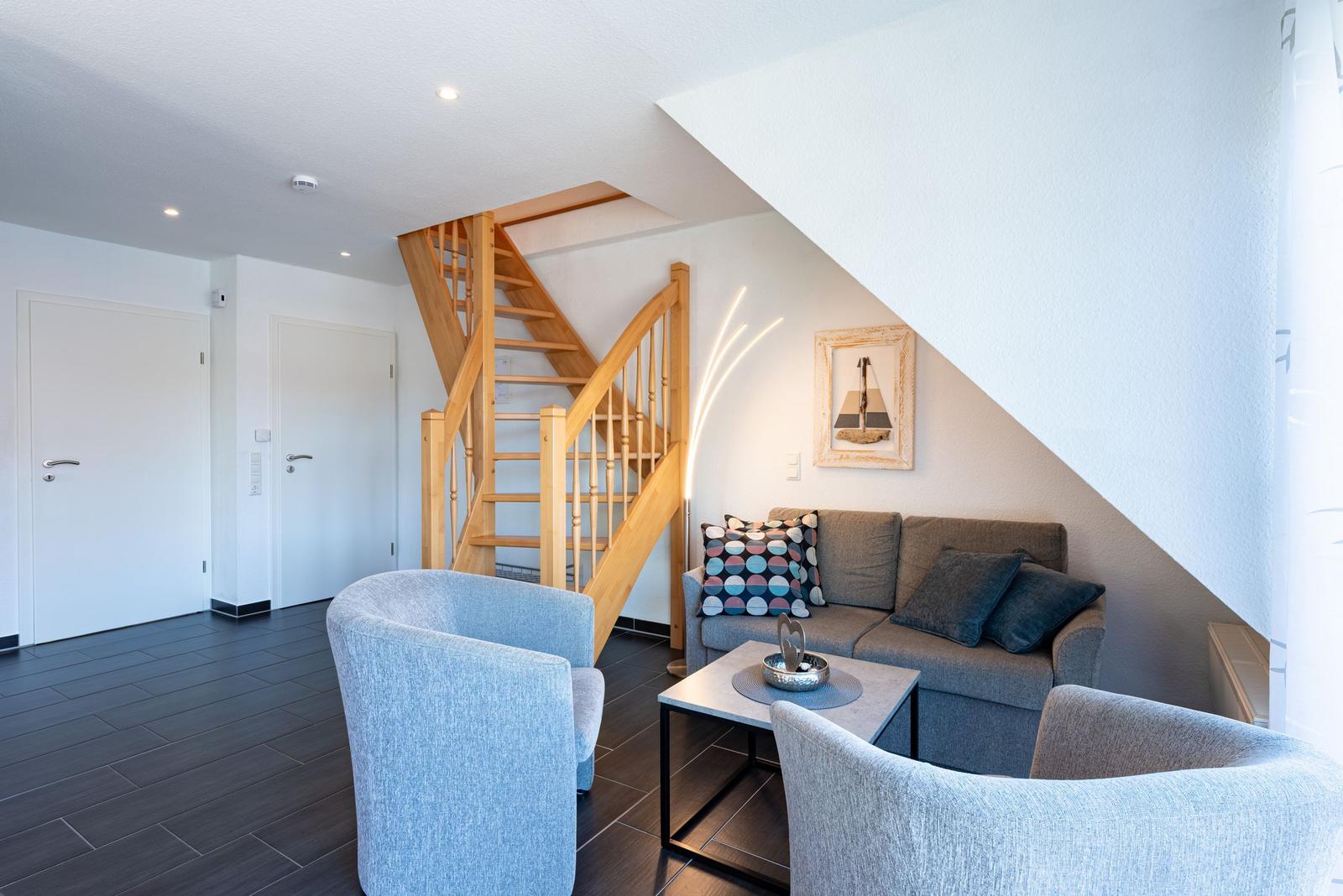 Wohnzimmer mit Sofa, zwei Sesseln und Holztreppe zur oberen Etage.