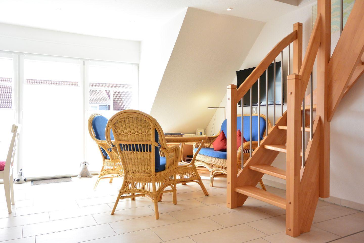 Wohnbereich mit Rattanmöbeln, Holztreppe und TV. Große Fenster mit Blick nach draußen.