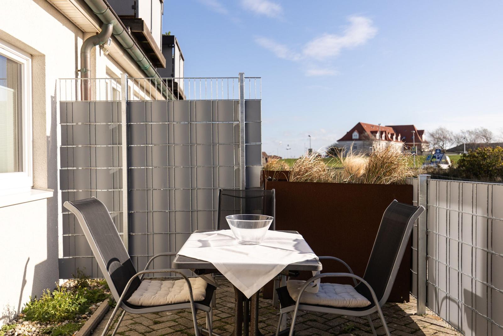 Terrasse mit Tisch und zwei Stühlen, umgeben von Zaun und Pflanzen.