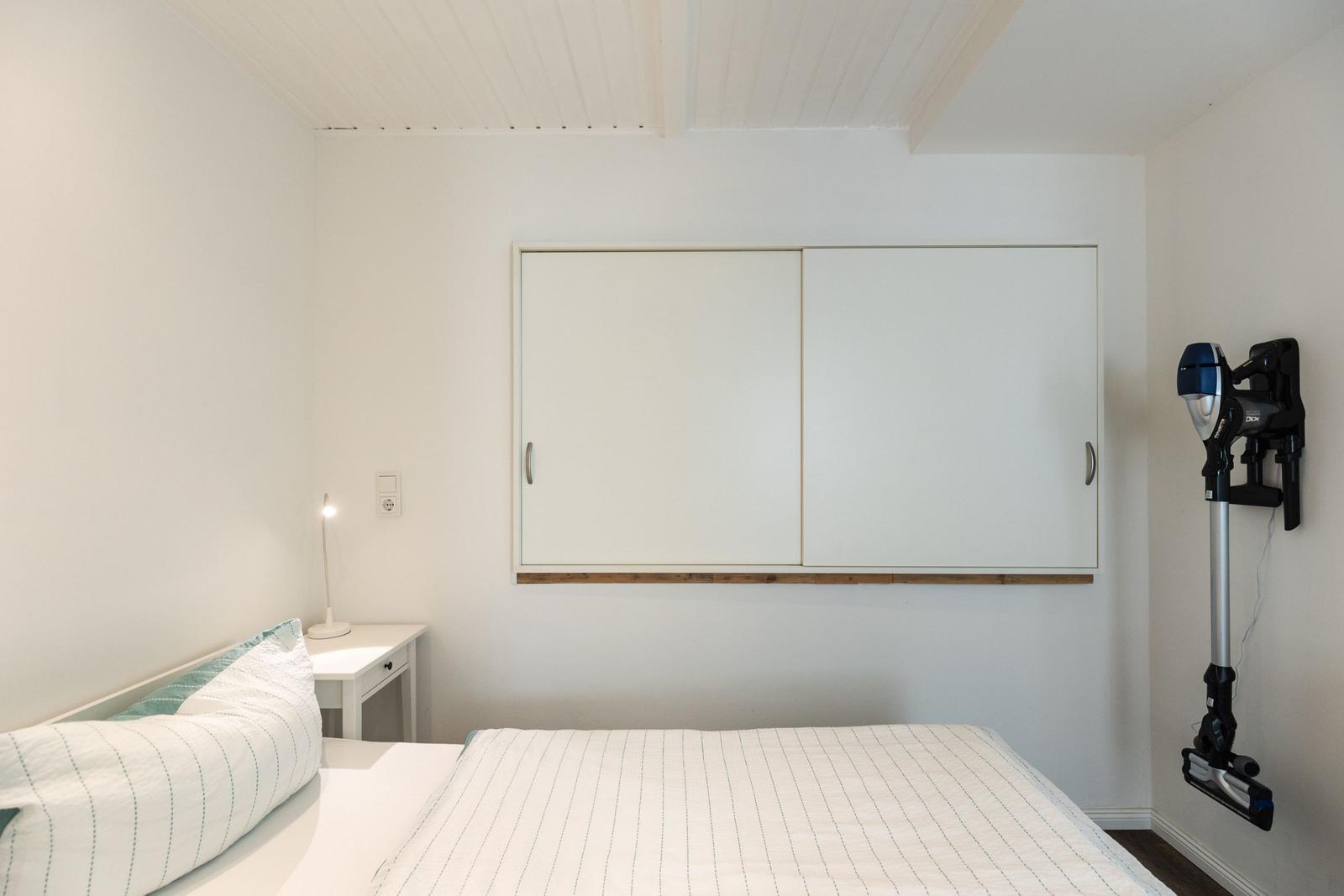 Schlafzimmer mit Bett, Nachttisch, Schrank und Staubsauger an der Wand.