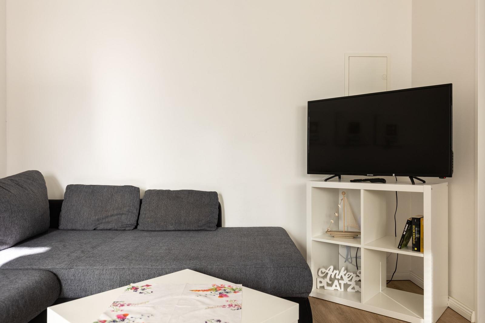 Ein grauer Sofa mit zwei Kissen, ein weißer Couchtisch und ein Fernseher auf einem weißen Regal.