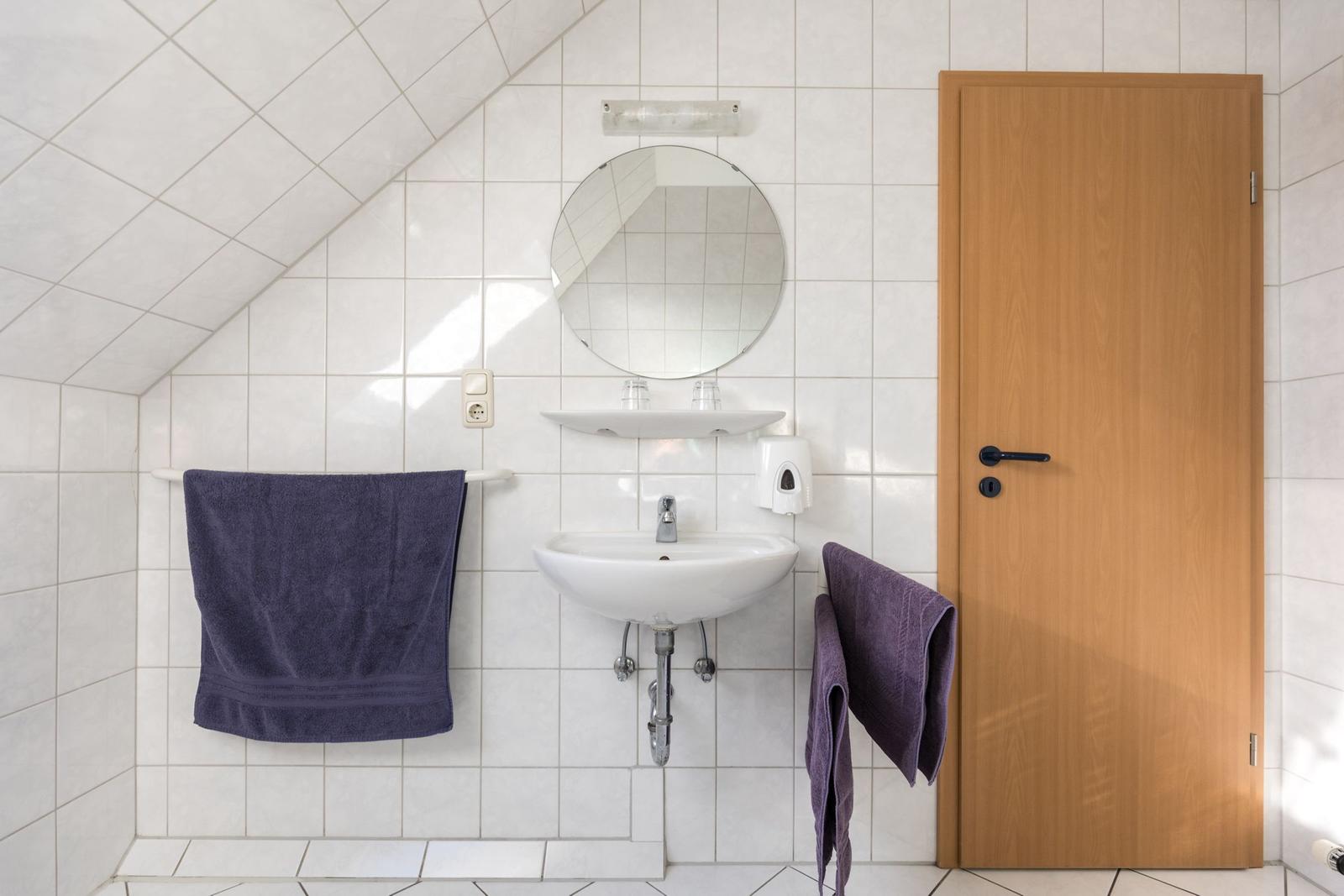 Badezimmer mit Waschbecken, Spiegel und Holztür. Dachneigung und Handtücher sichtbar.