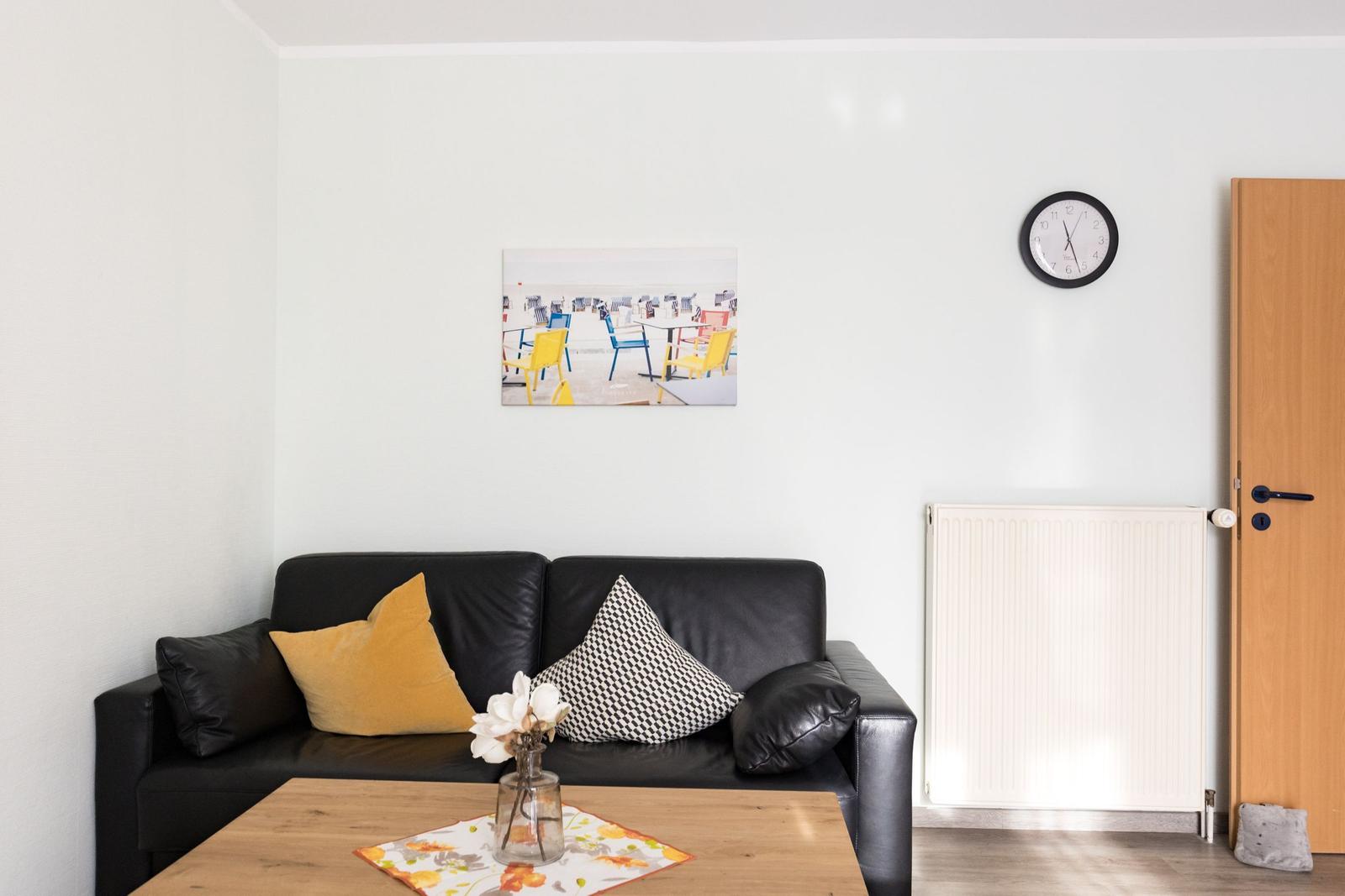 Schwarzes Sofa mit Kissen, Tisch, Wanduhr und Bild an der Wand.