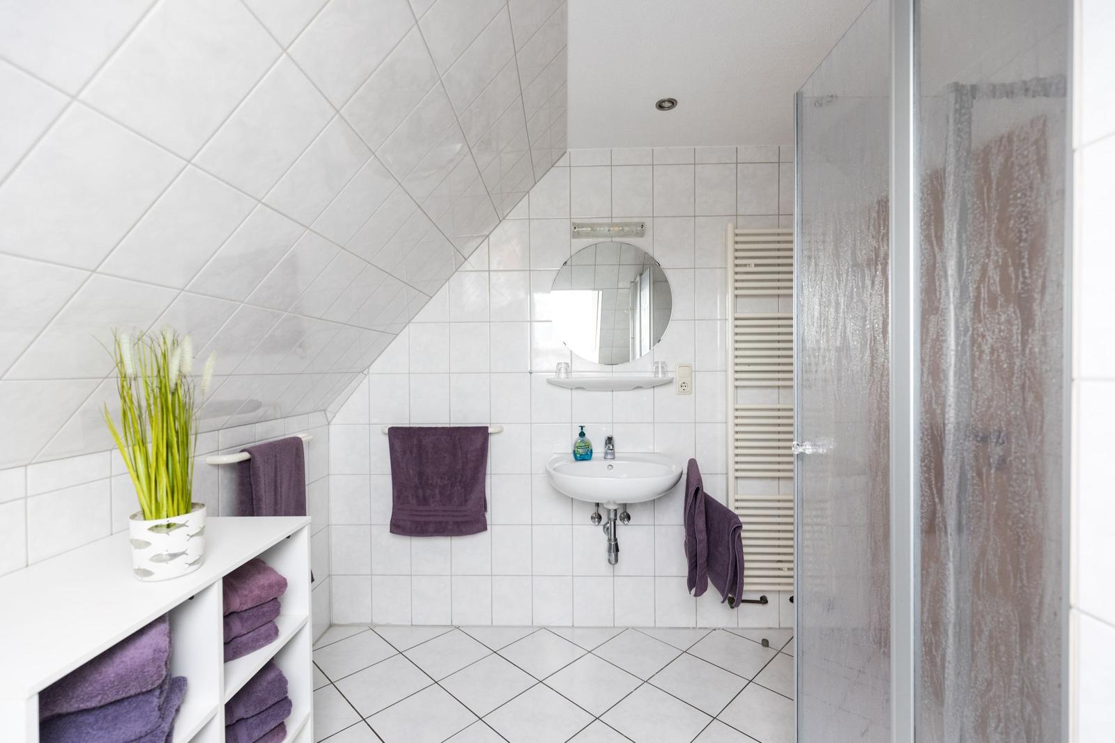 Badezimmer mit Dusche, Waschbecken und Dachneigung. Lila Handtücher und Pflanze.