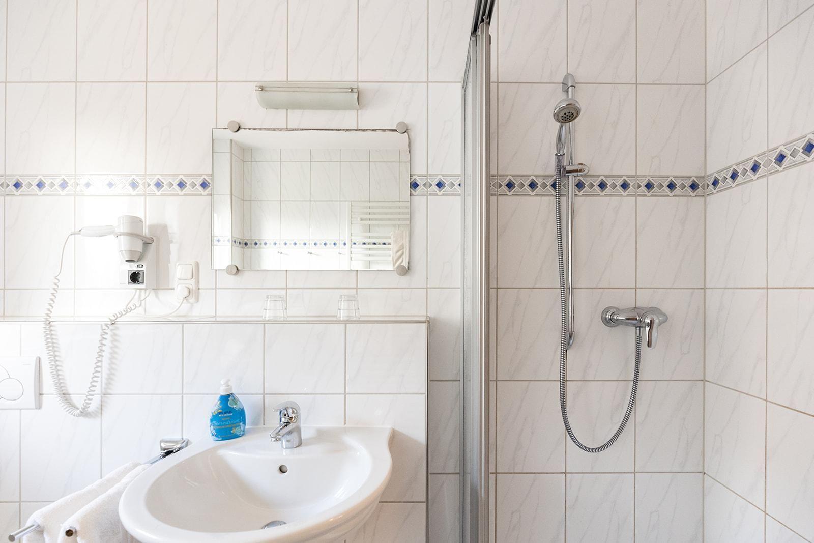 Badezimmer mit Dusche, Waschbecken und Spiegel. Weiße Fliesen mit blauer Zierleiste.