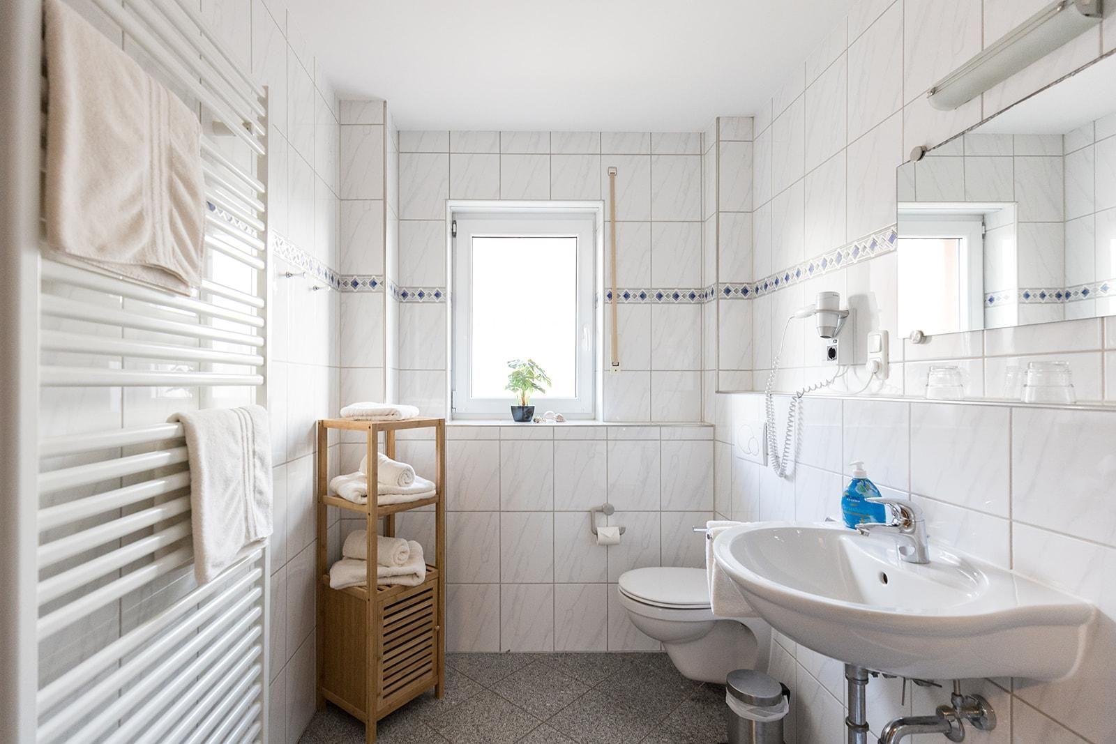 Badezimmer mit Toilette, Waschbecken und Heizung. Holzregal und Fenster mit Pflanze.