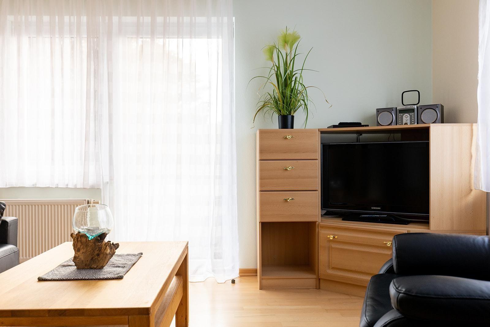 Wohnzimmer mit Holzmöbeln, Fernseher und Pflanze. Tisch mit Glasobjekt und Fenster mit Vorhängen.
