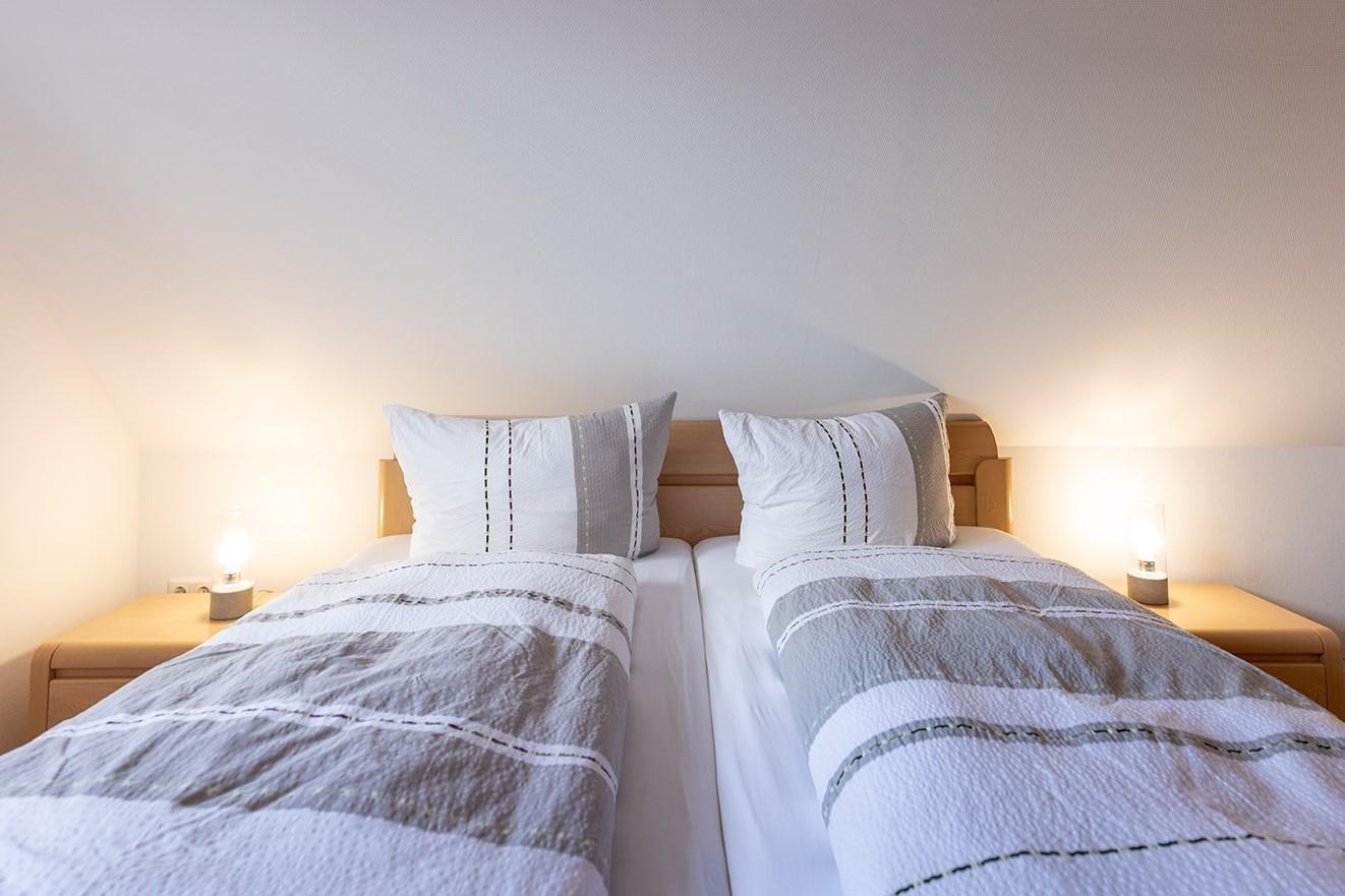 Zwei Betten mit Kissen und Bettwäsche unter einer schrägen Decke mit Nachttischen und Lampen.
