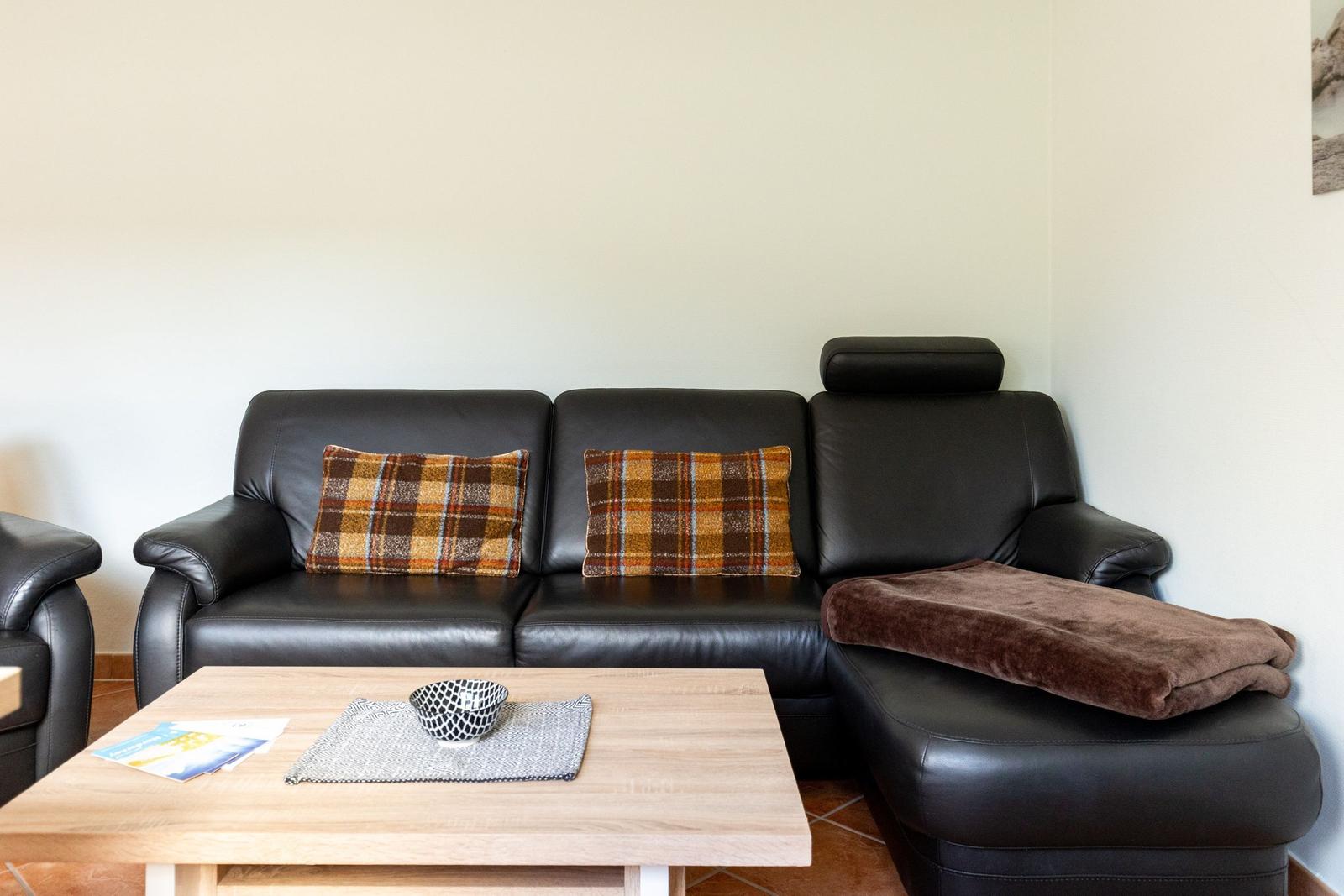 Schwarzes Ledersofa mit karierten Kissen und brauner Decke. Holztisch mit Schale und Broschüren.