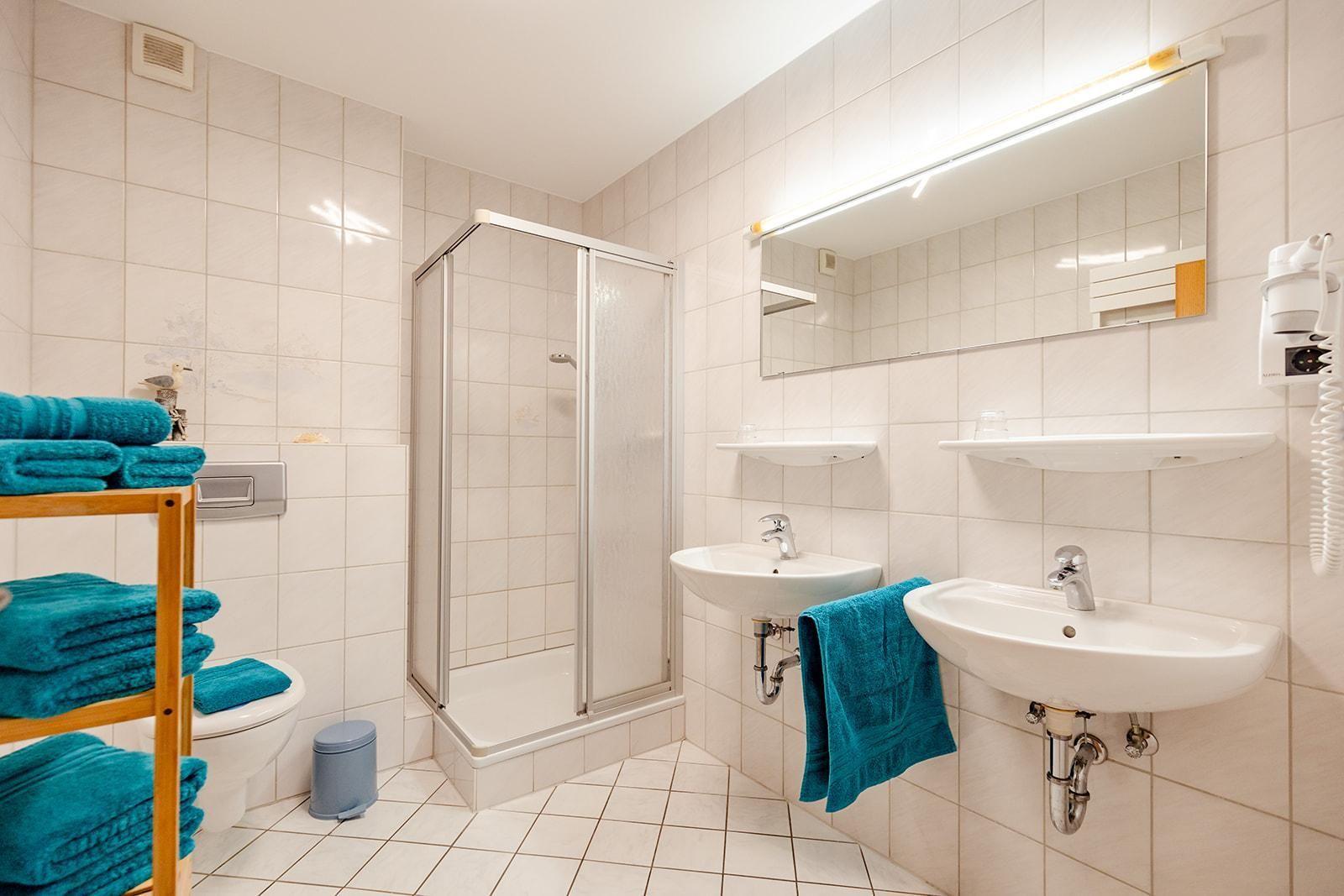 Badezimmer mit Dusche, zwei Waschbecken und Toilette. Blaue Handtücher und Spiegelschrank.