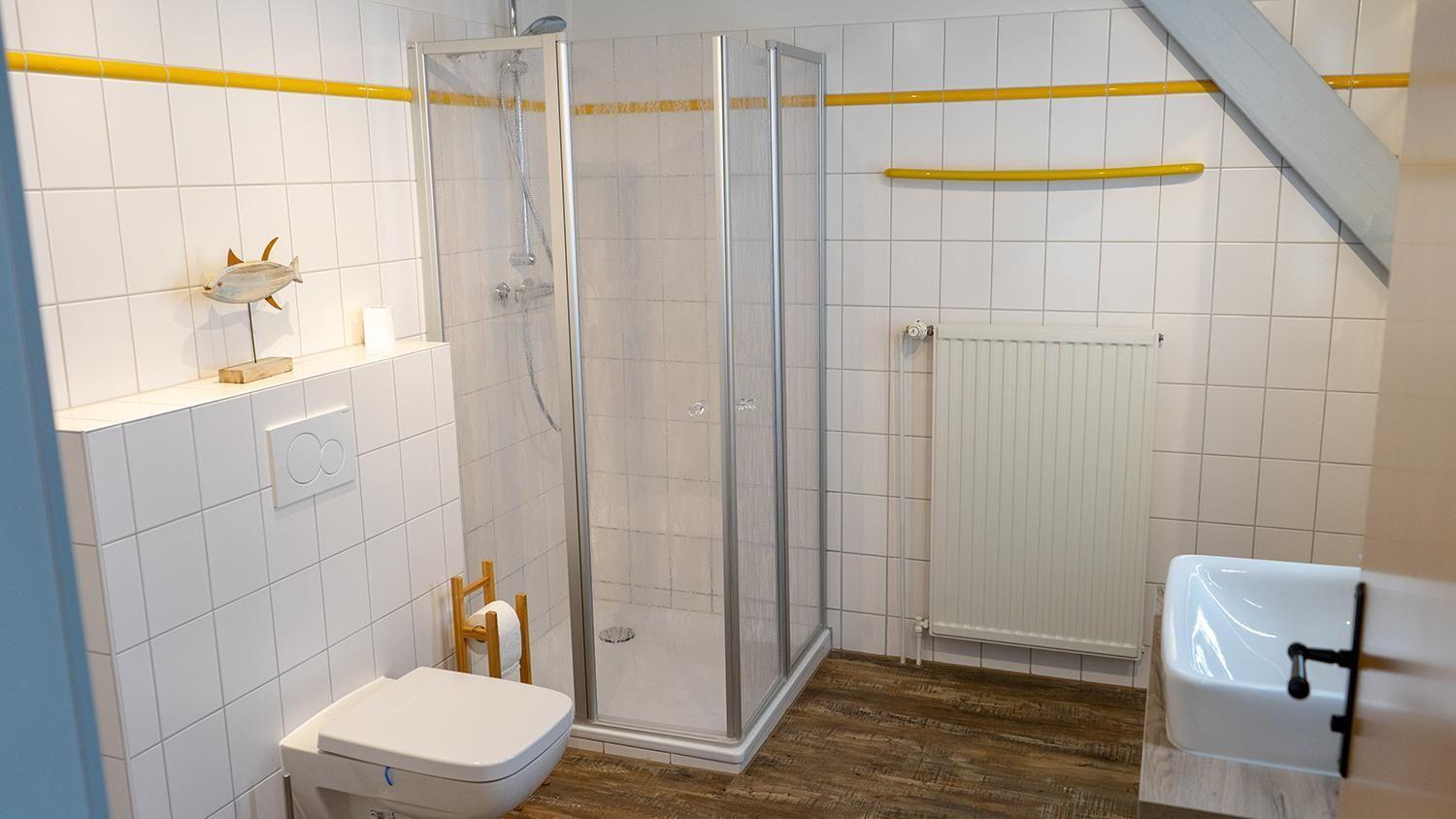 Bathroom with shower, toilet, and sink. White tiles and wooden floor.
