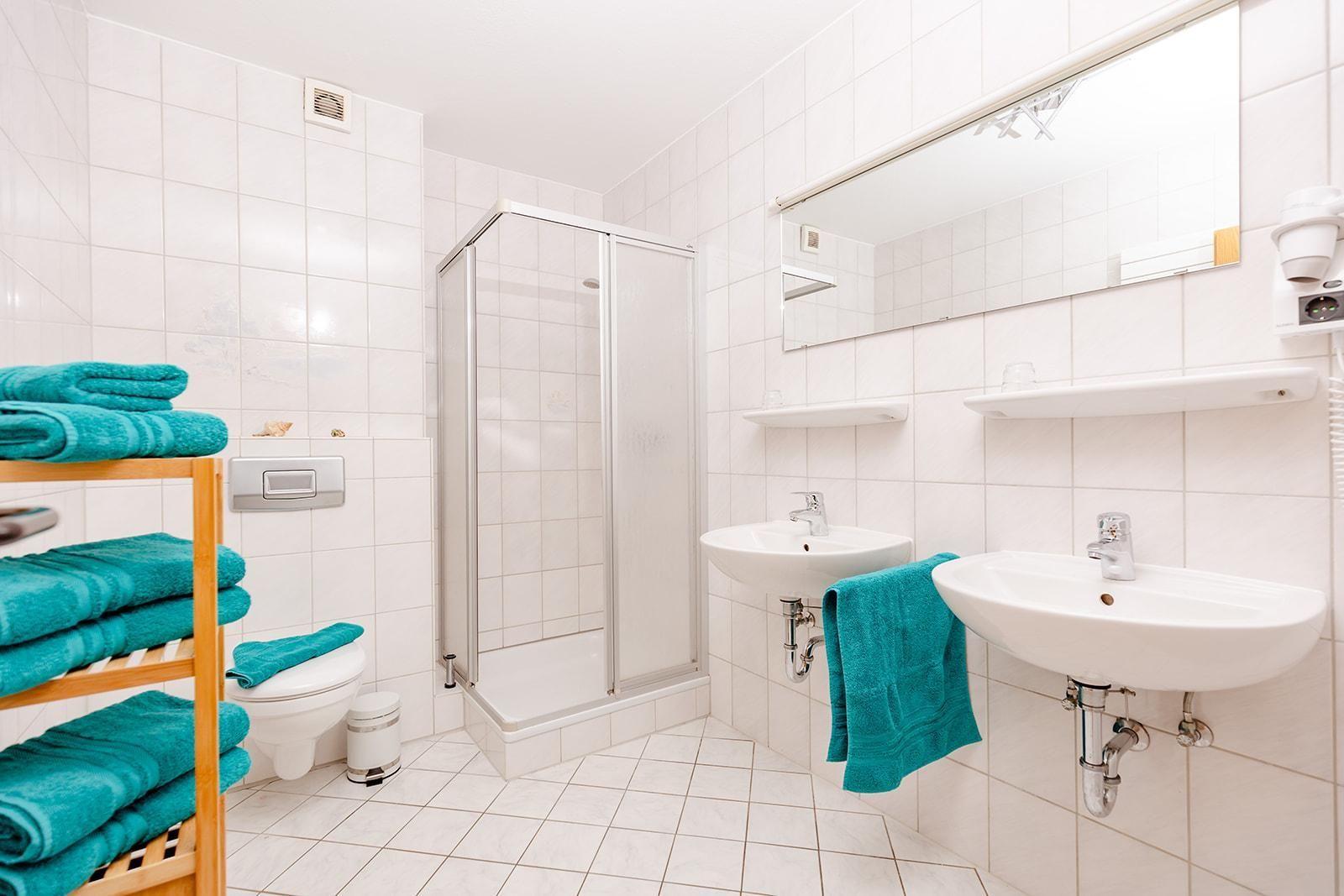 Badezimmer mit Dusche, zwei Waschbecken und Toilette. Türkis Handtücher und Spiegelschrank.