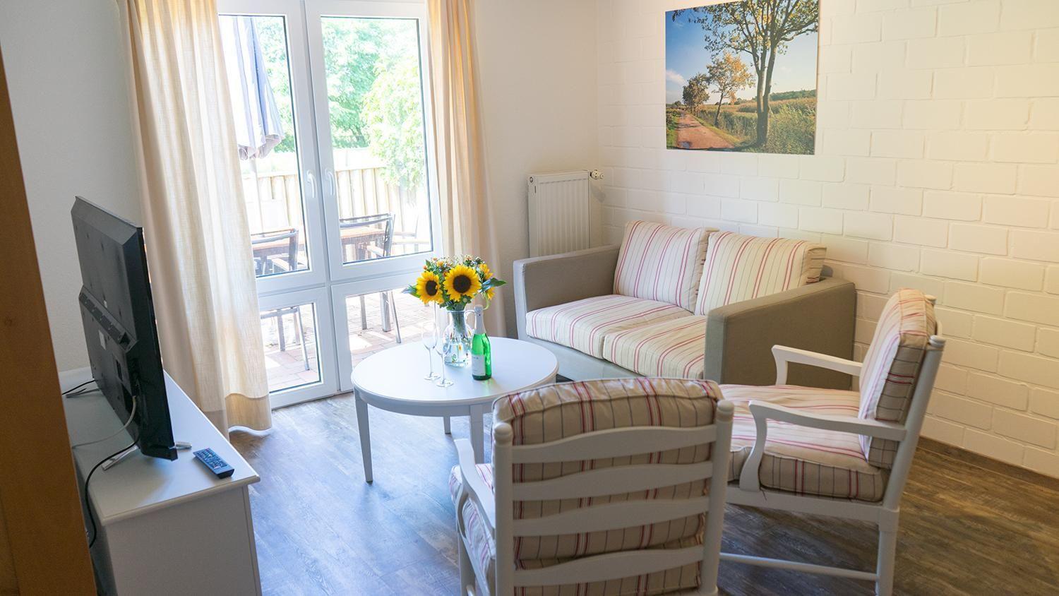 Wohnzimmer mit Sofa, zwei Stühlen, Tisch und Fernseher. Durchgang zur Terrasse.