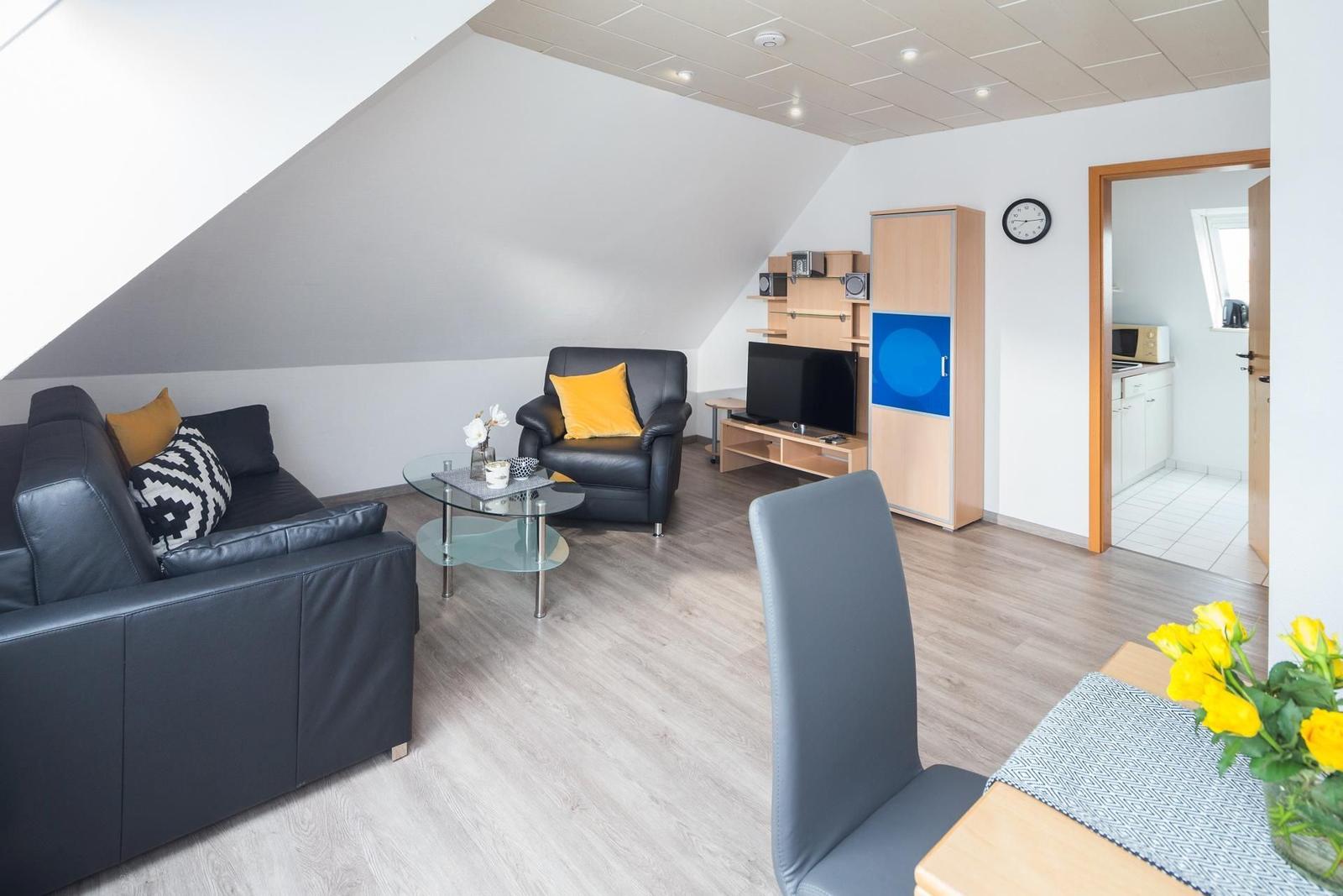 Wohnraum mit Sofa, Sessel, Fernseher und Küchenbereich im Hintergrund.