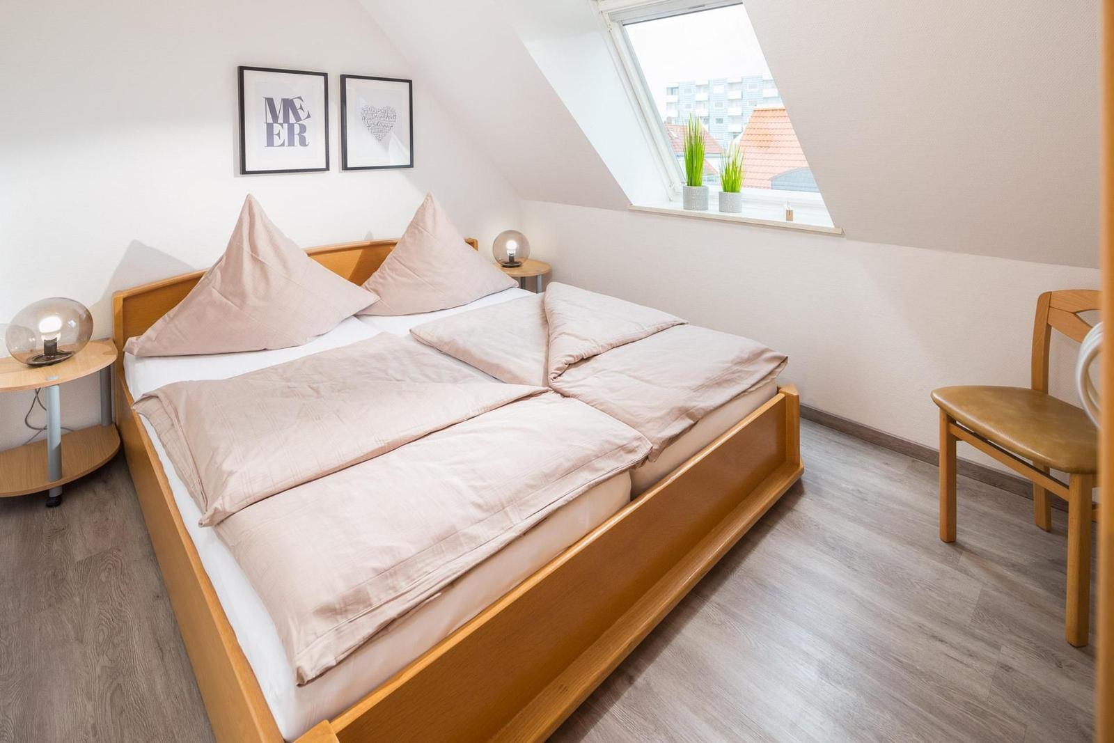 Schlafzimmer mit Dachschrägen, Bett, Nachttisch und Stuhl. Fenster mit Blick auf Dächer.