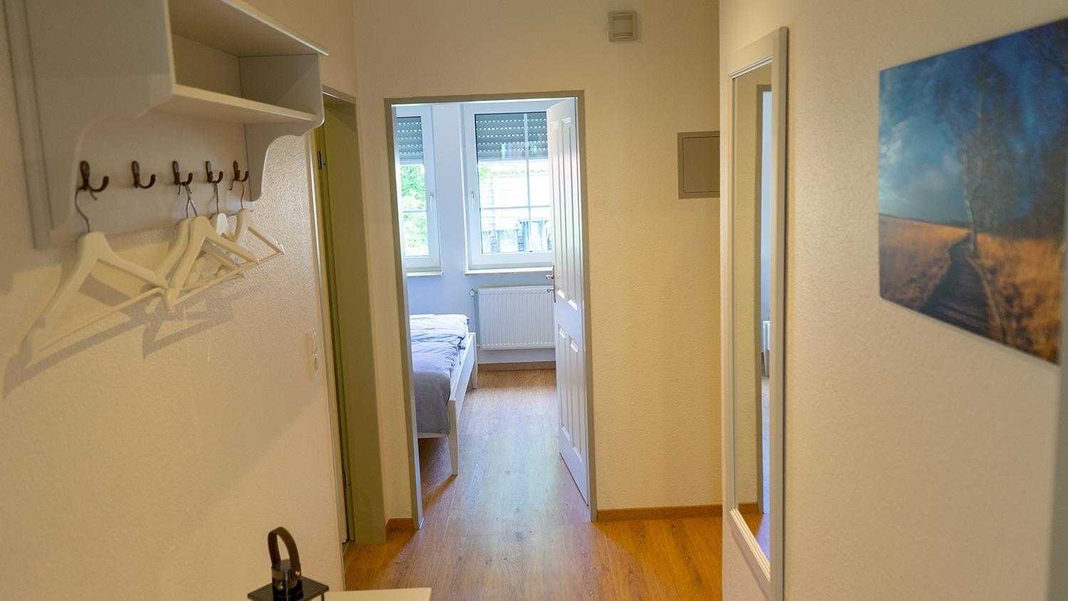 Flur mit Kleiderhaken, Spiegel und Bild an der Wand. Im Hintergrund ist ein Schlafzimmer mit Bett und Fenster.