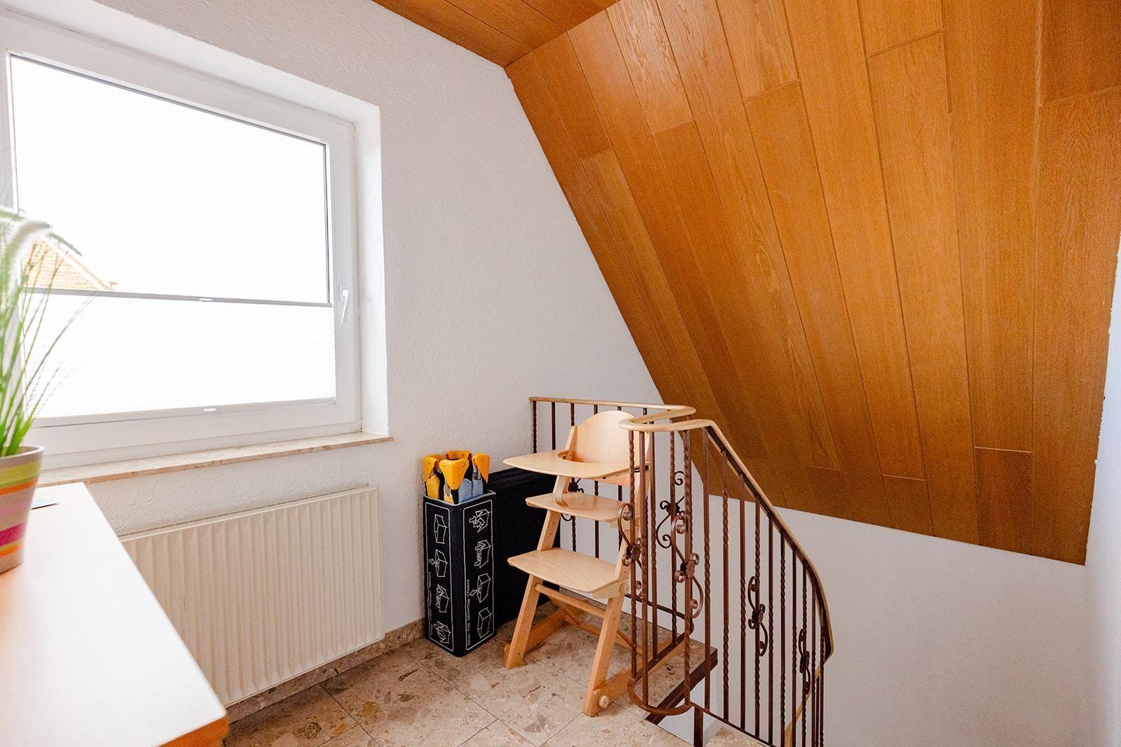 Dachgeschoss mit Holzdecke, Stiege und Fenster. Kinderhochstuhl und Pflanze sichtbar.