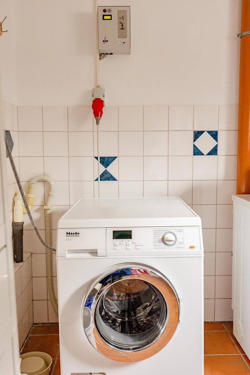 Waschmaschine in einem Raum mit Fliesenwand und rotem Wasserhahn.