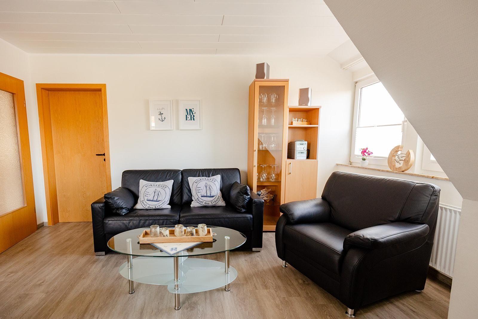 Wohnzimmer mit schwarzer Ledercouch, Glas-Tisch und Holzmöbeln unter Dachschrägen.