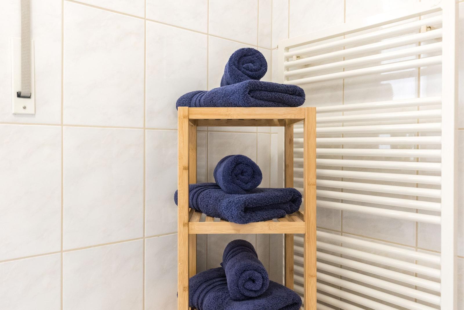 Drei blaue Handtücher auf einem Holzregal in einem Badezimmer mit weißen Fliesen.