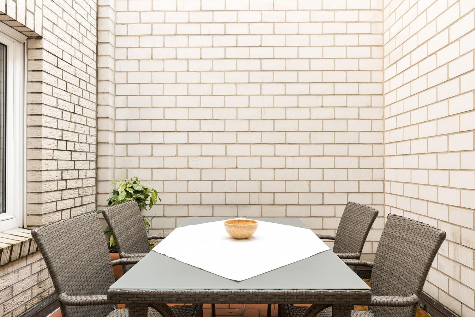 Terrasse mit Tisch und vier Stühlen, weiße Ziegelwand im Hintergrund.