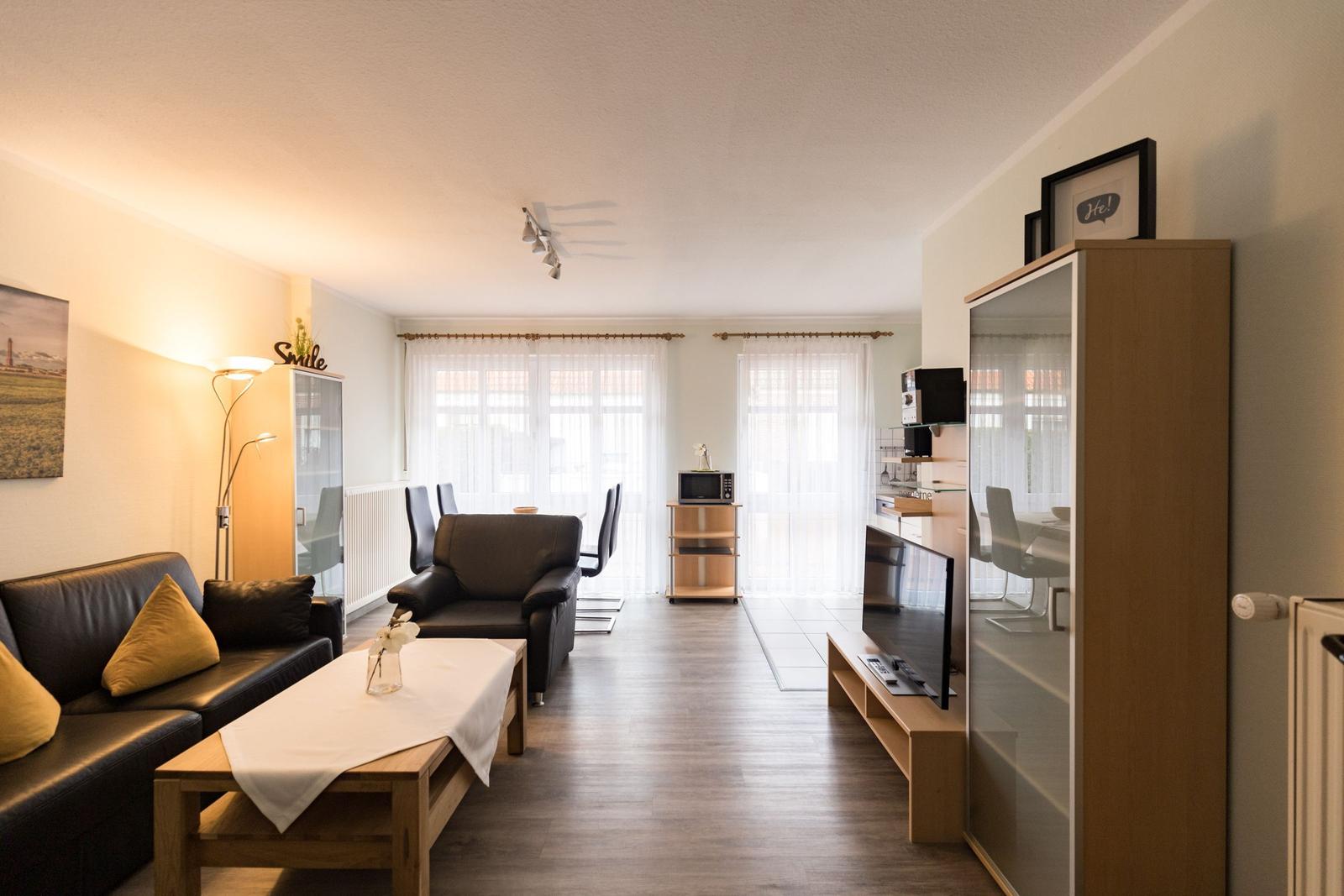 Wohnzimmer mit Sofa, Sessel, Couchtisch und Fernseher. Hintergrund: Essbereich mit Tisch und Stühlen.