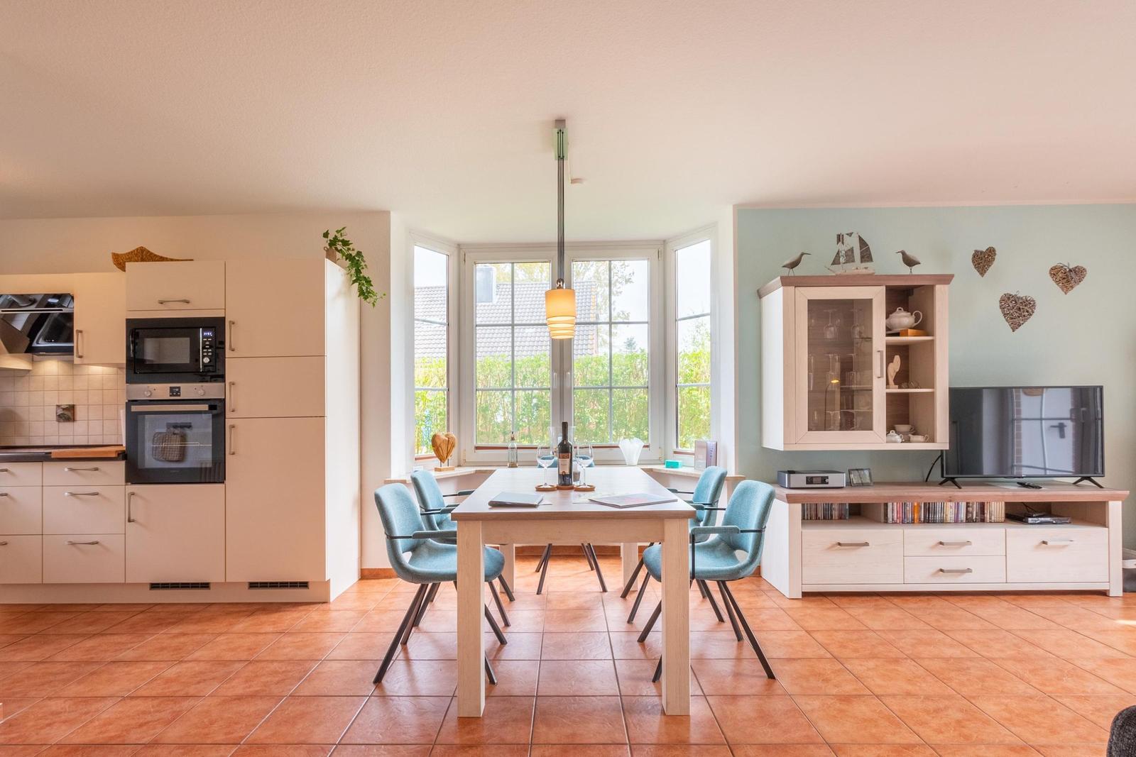 Küche mit Essbereich und TV-Ecke. Holztisch, blaue Stühle, Fenster mit Blick nach draußen.