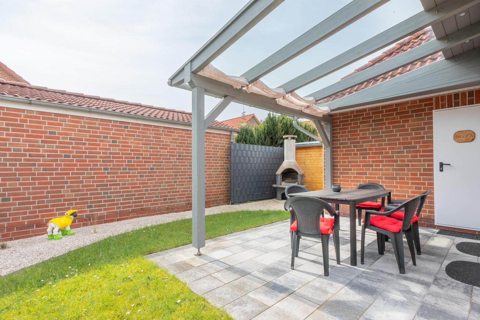 Terrasse mit Tisch, Stühlen und Grill unter Pavillon. Grünfläche und Mauer im Hintergrund.