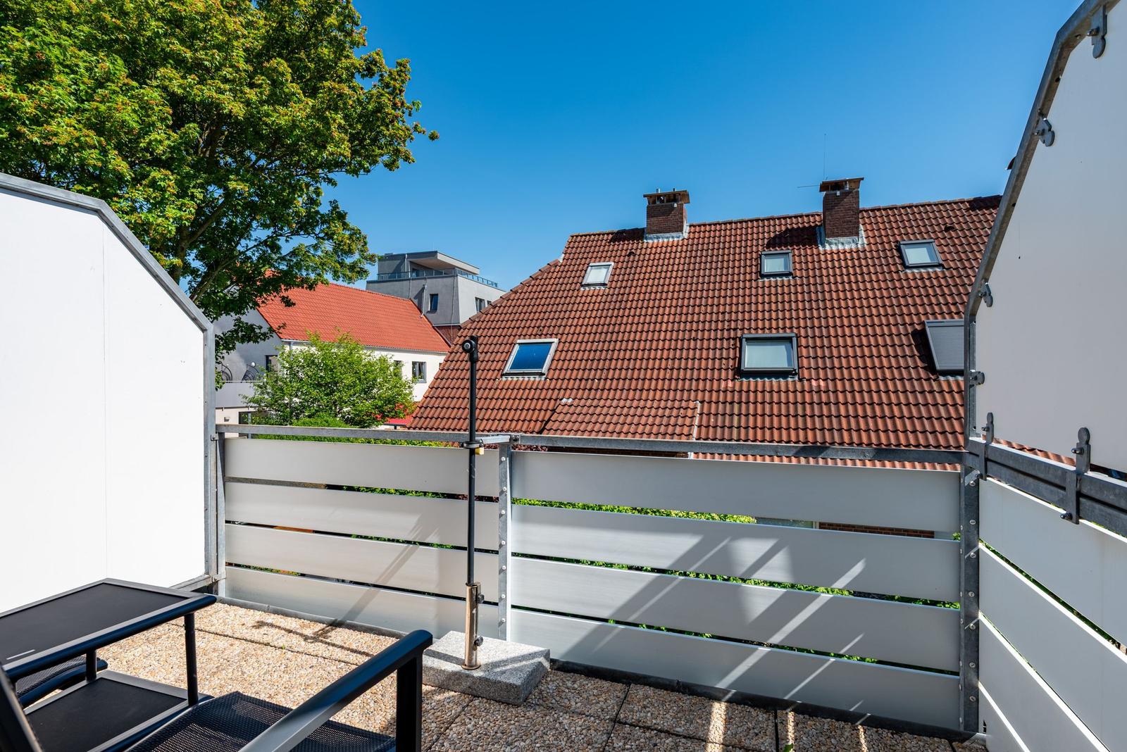 Terrasse mit Gartenzaun und Blick auf Dach mit Schornsteinen und Fenstern.