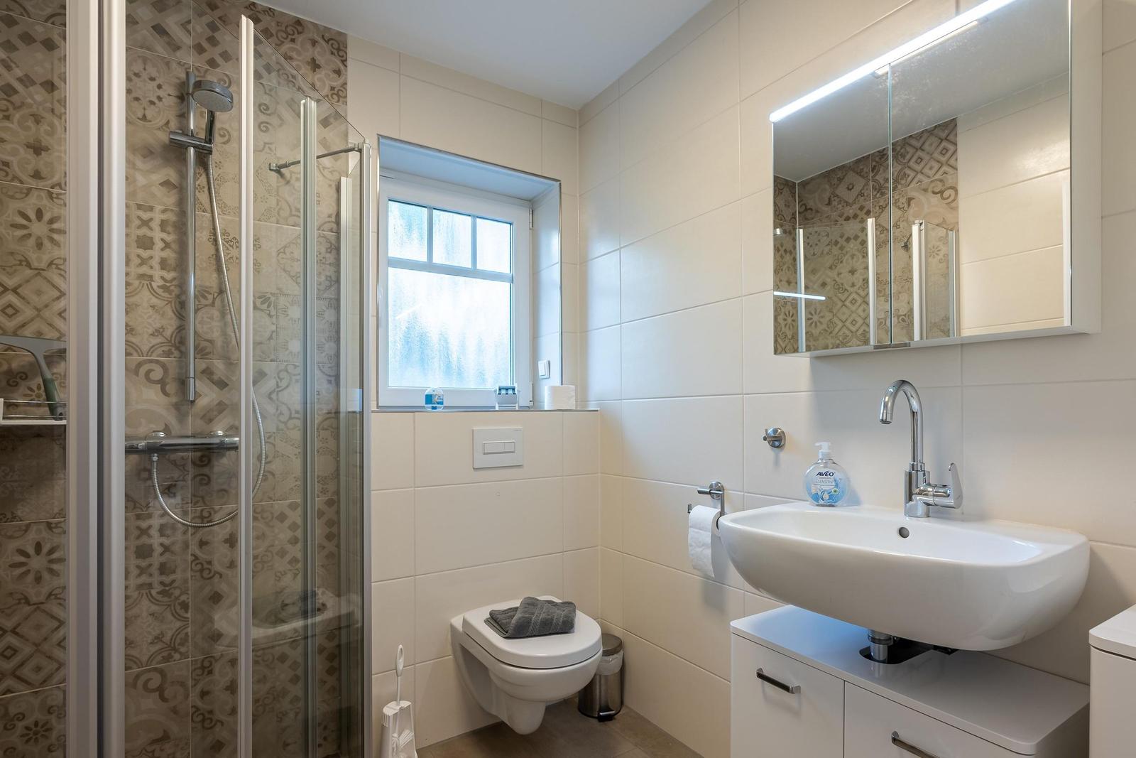 Badezimmer mit Dusche, Waschbecken und Toilette. Fenster sorgt für Tageslicht.