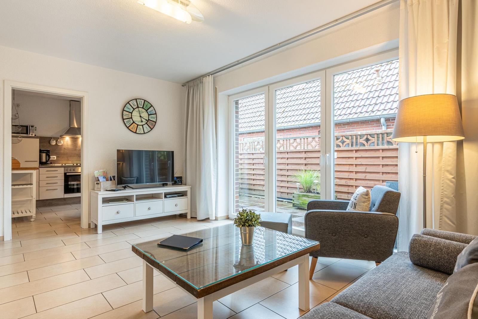 Wohnzimmer mit Glas-Tisch, Sofa und Sessel. Durchgang zur Küche. Fenster zur Terrasse.