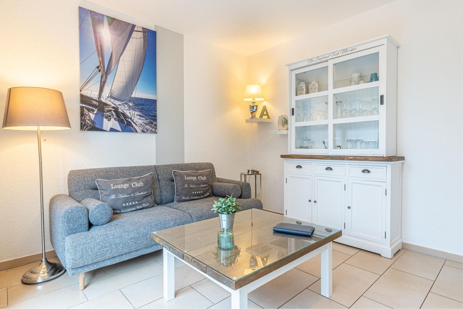 Ein Wohnzimmer mit grauem Sofa, Glas-Tisch und weißem Schrank. Wandbild zeigt Segelboot.