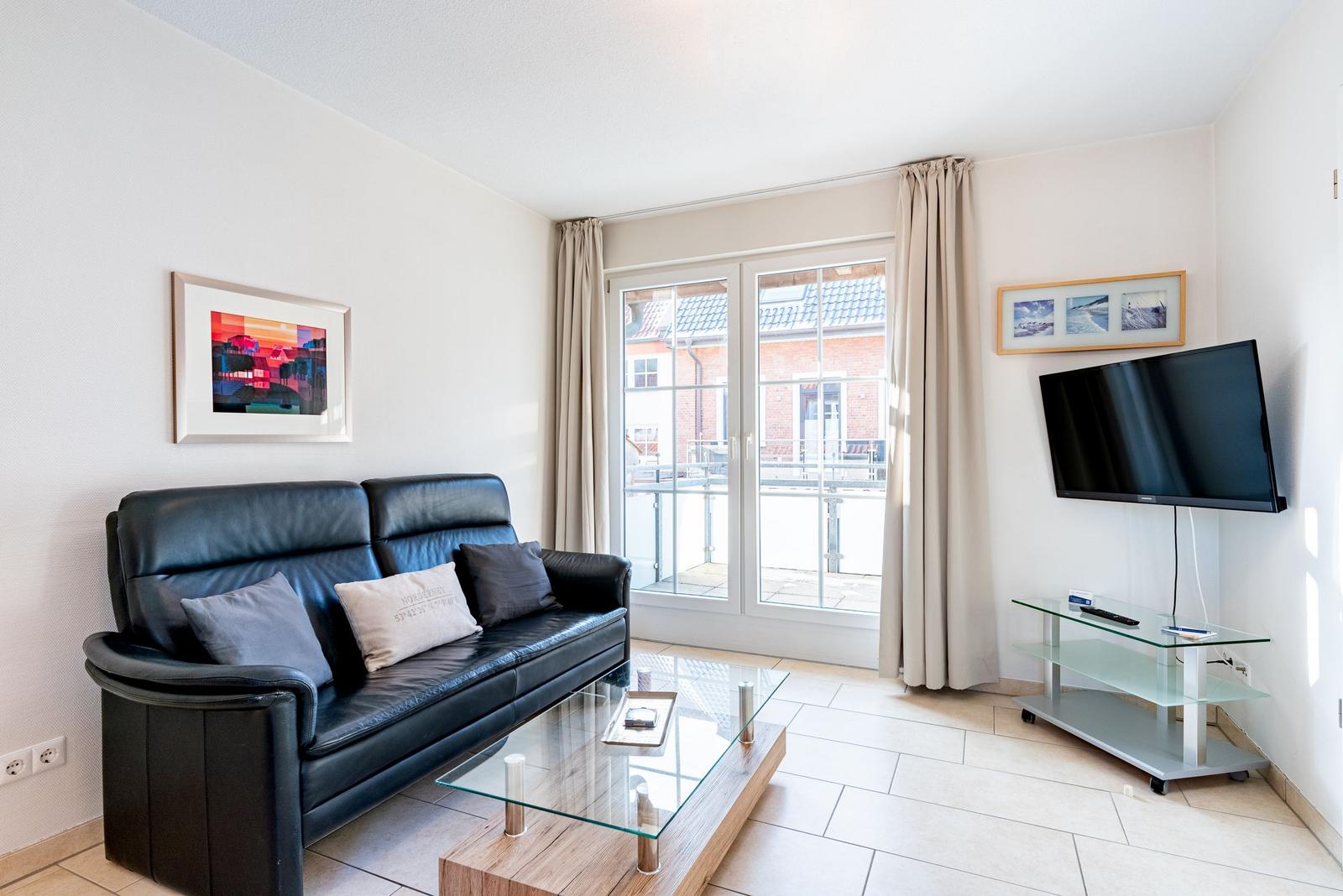 Wohnzimmer mit Sofa, Glas-Tisch und Fernseher. Durchgangstür zum Balkon.