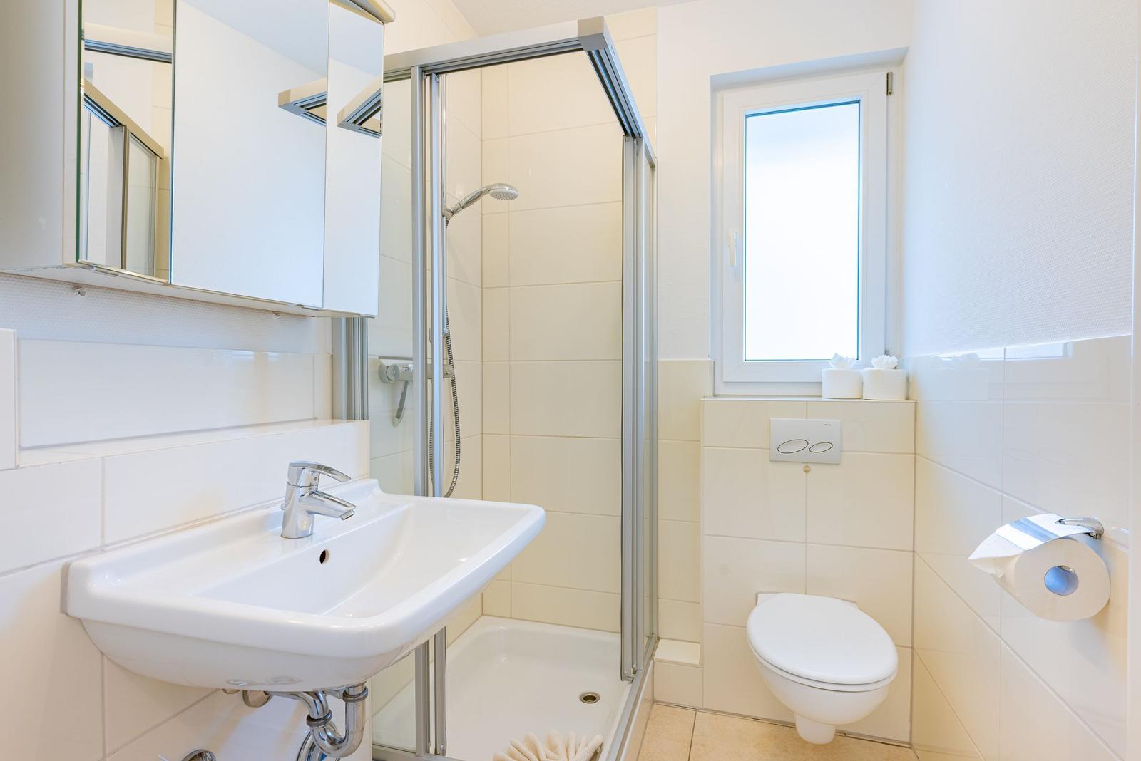 Badezimmer mit Dusche, Waschbecken und WC. Fenster sorgt für Tageslicht.