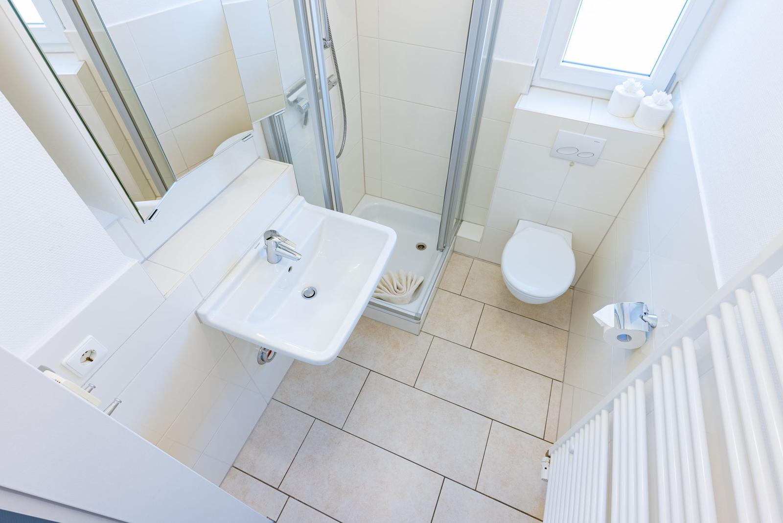 Badezimmer mit Dusche, Waschbecken und Toilette. Fenster und Heizung sind sichtbar.