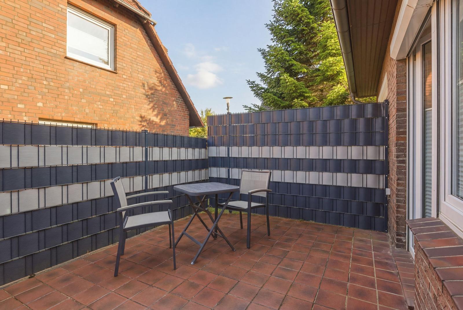 Terrasse mit Tisch und zwei Stühlen, umgeben von einem Zaun und einem Haus.