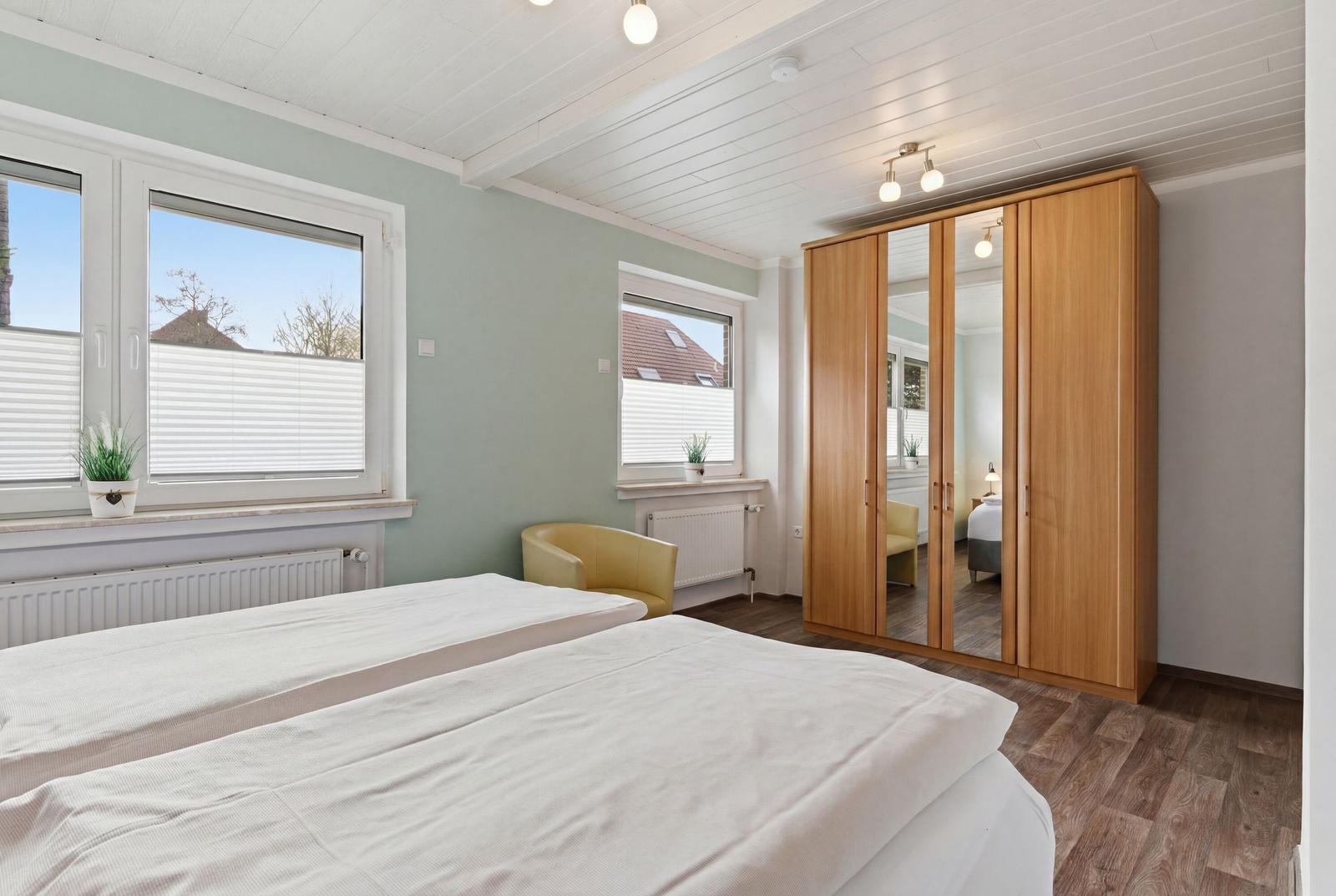 Schlafzimmer mit Doppelbett, Holzschrank und Fenstern mit Jalousien.