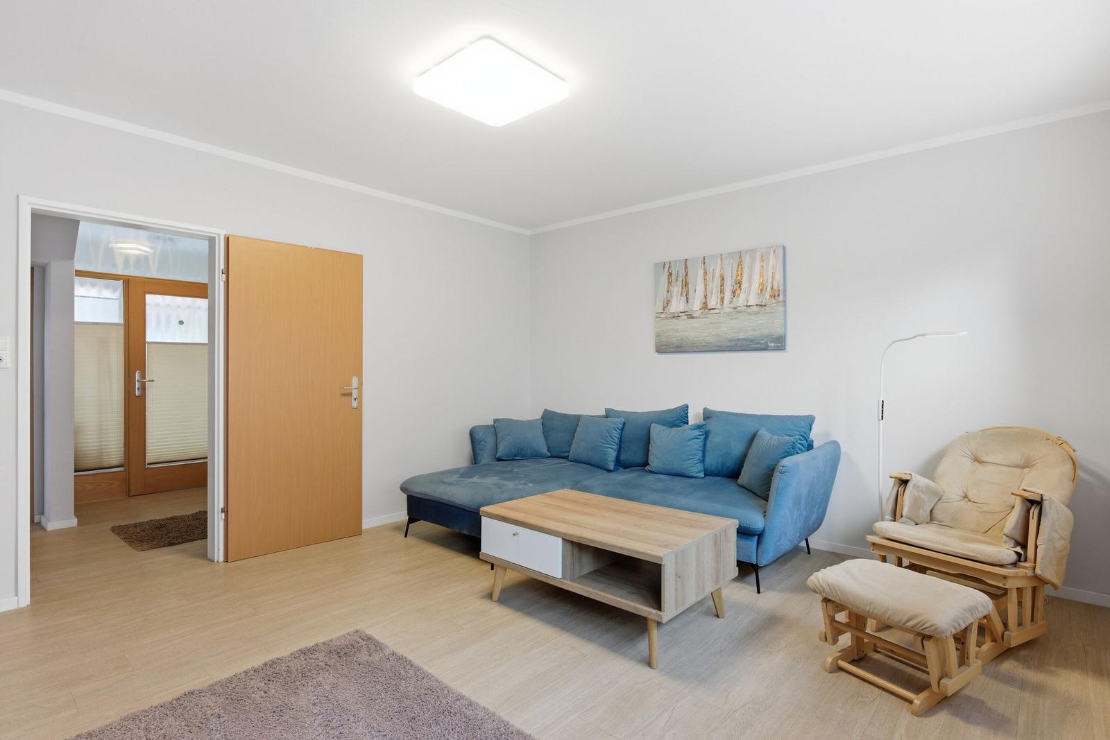 Wohnzimmer mit blauem Sofa, Holztisch und Schaukelstuhl. An der Wand hängt ein Bild.