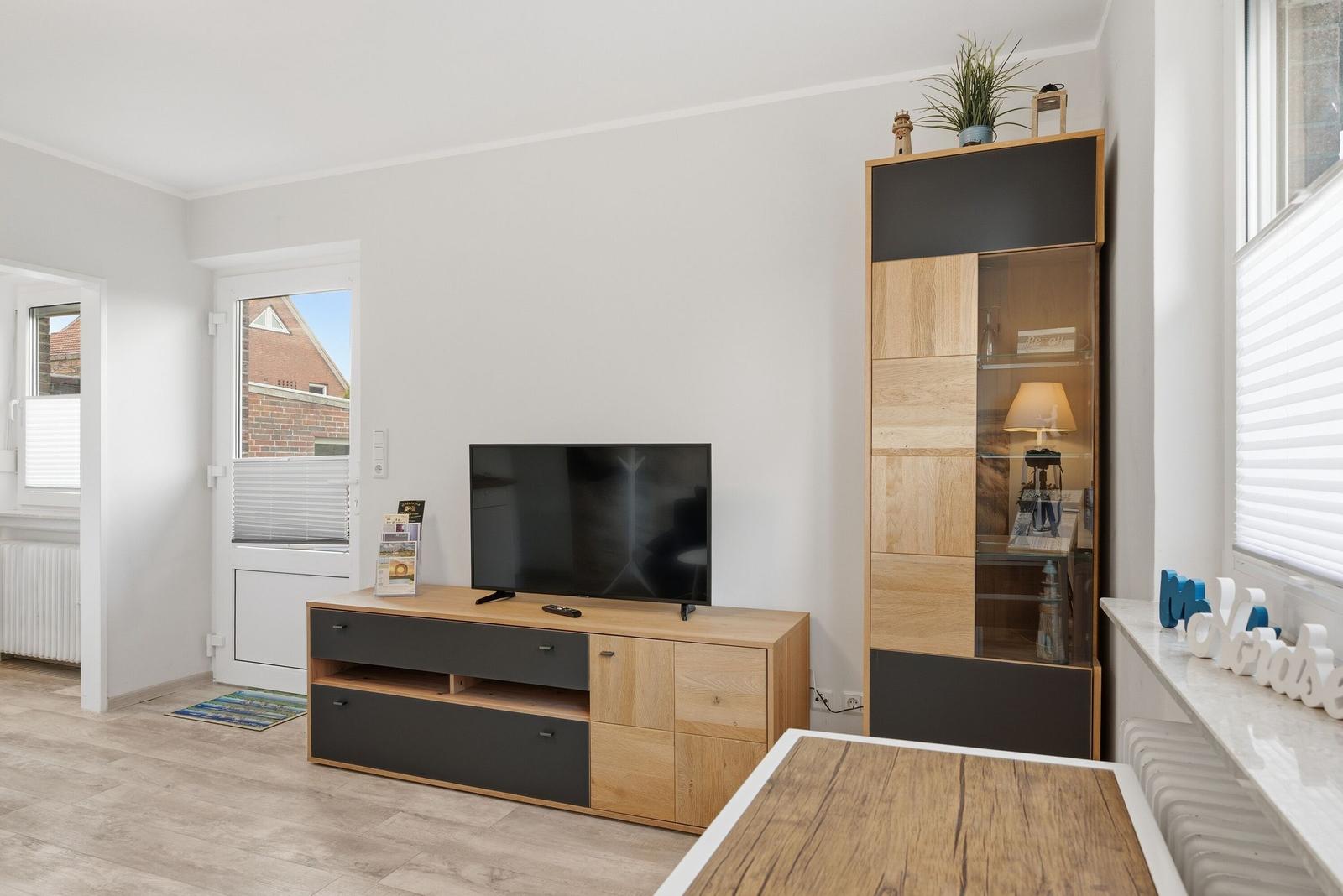 Wohnzimmer mit Fernseher, Schrank und Fenster. Holzmöbel und helle Wände.
