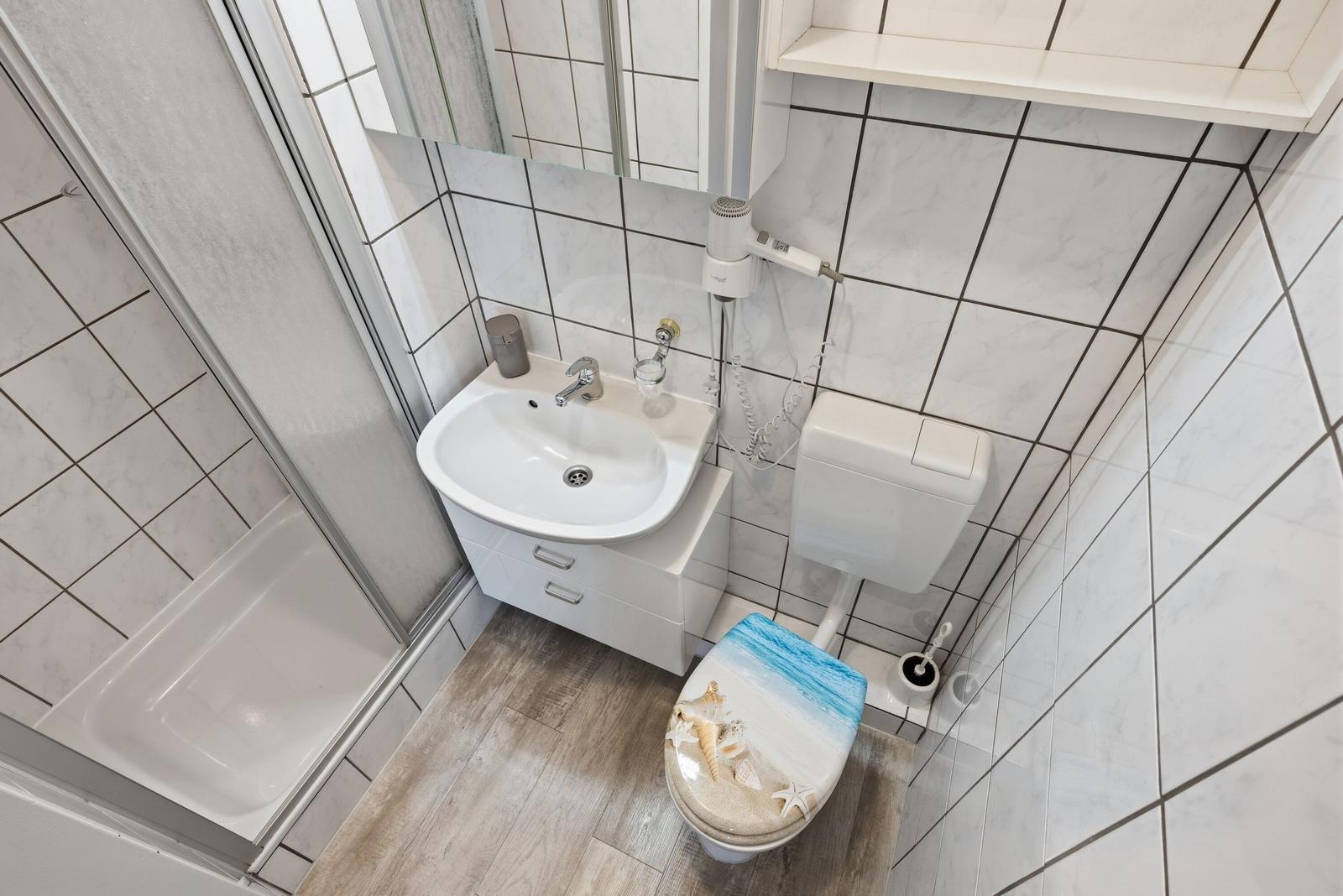 Badezimmer mit Dusche, Waschbecken und Toilette mit Strandmotivdeckel.